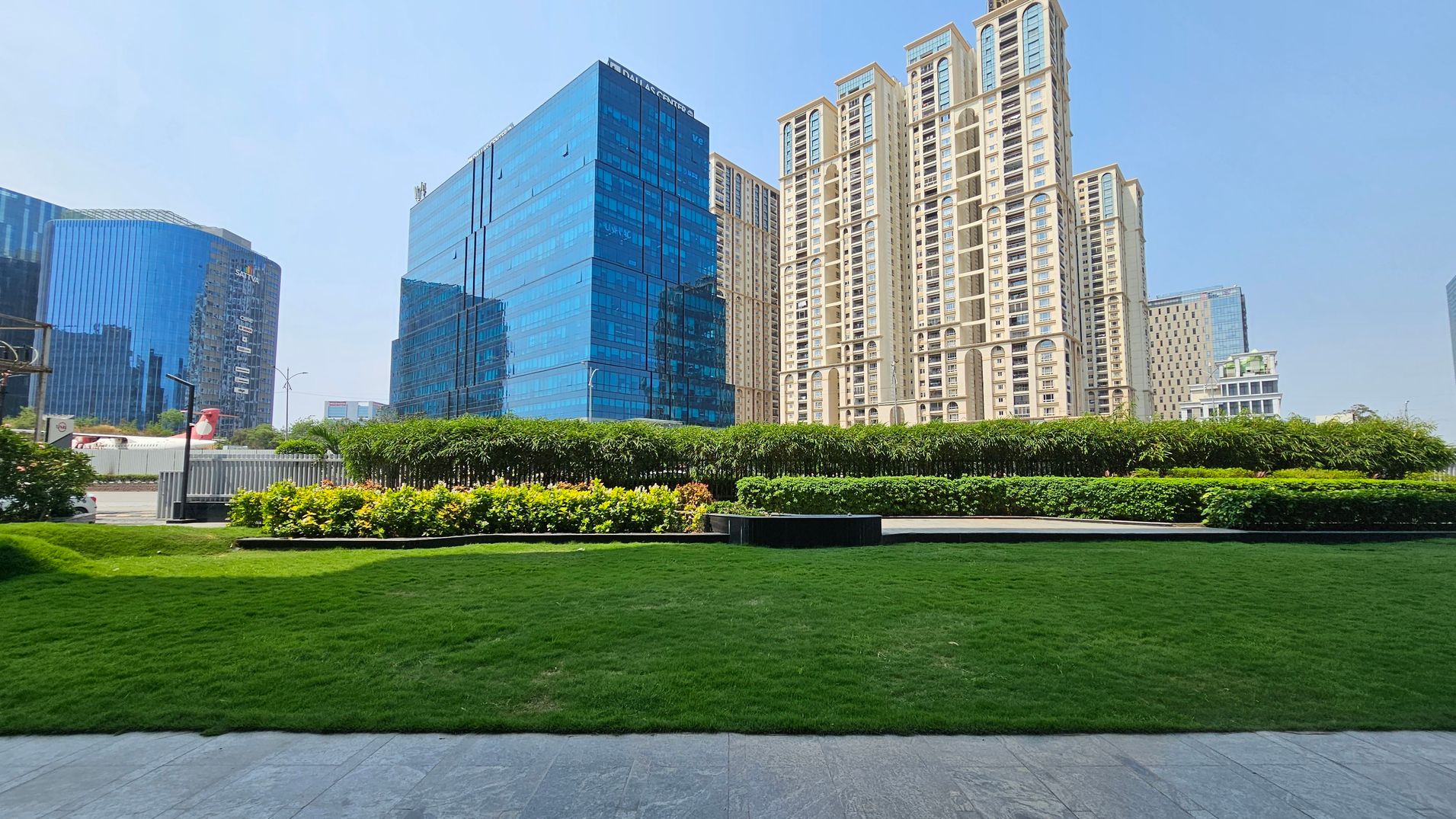 In the foreground, a well-maintained green lawn is bordered by a stone pathway. The middle ground features a row of neatly trimmed bushes and small flowering plants. In the background, several tall buildings with glass facades and modern architecture rise against a clear sky. The buildings vary in design, with some having reflective blue glass and others a beige exterior with multiple windows.