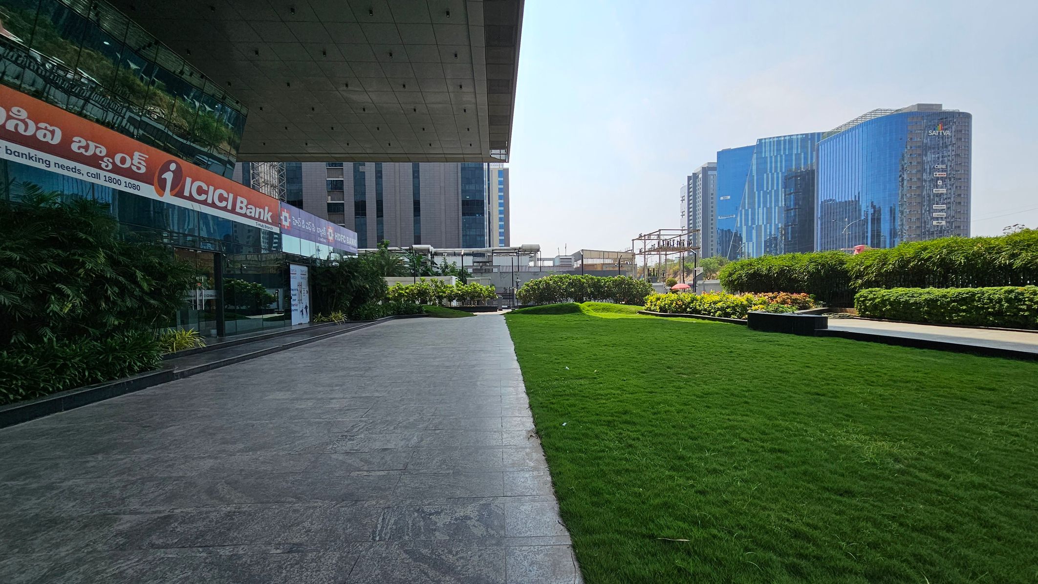 On the left, a building with reflective glass walls displays signage for ICICI Bank. The foreground features a paved walkway adjacent to a well-maintained green lawn. In the background, modern high-rise buildings with blue-tinted glass facades are visible. The right side is lined with neatly trimmed hedges and flowering plants. The ceiling of the building overhangs slightly above the walkway.