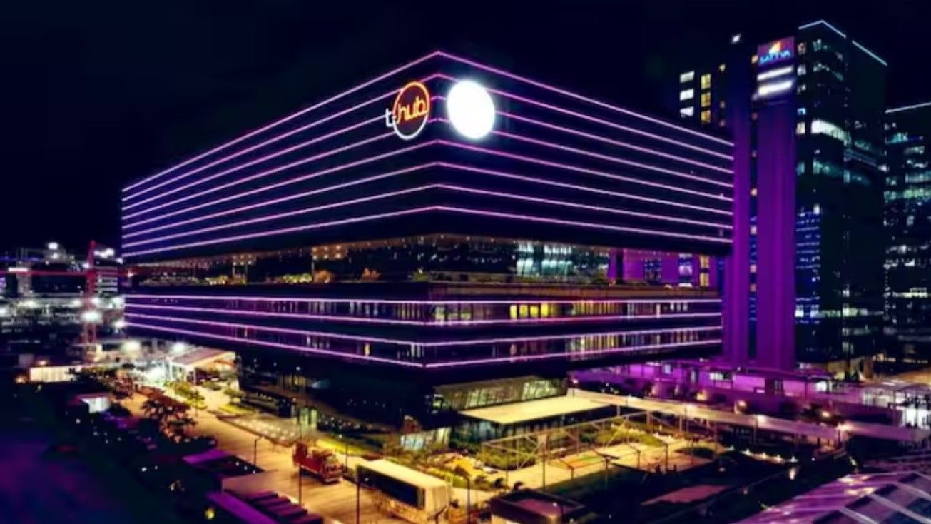 A modern building at night with horizontal lines of purple and pink lights across its facade. The structure is large and rectangular, with a circular logo on the upper section. Surrounding the building are other illuminated high-rise structures. The foreground features a well-lit area with pathways and parked vehicles. The overall scene is vibrant with artificial lighting.