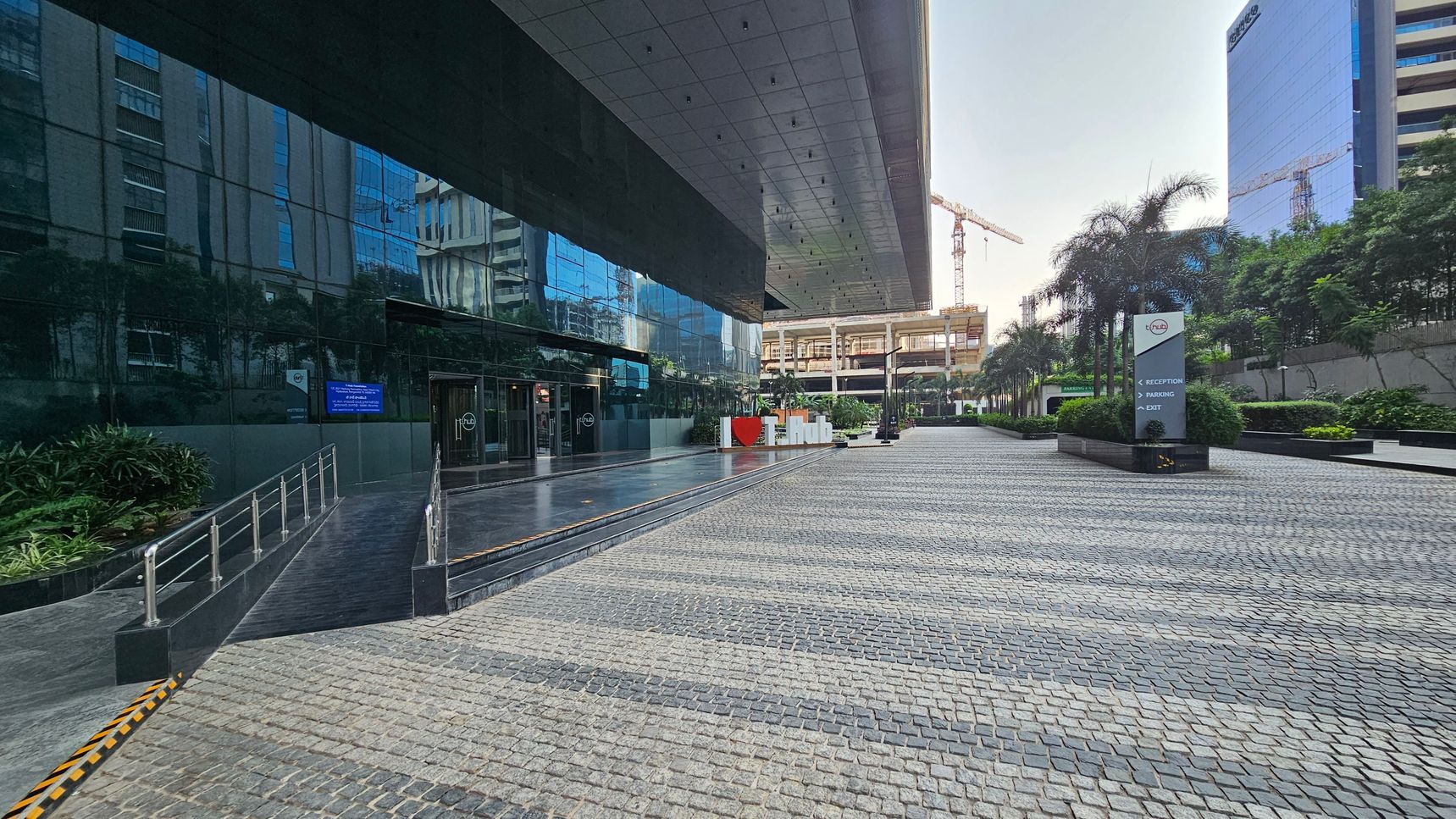 A modern building with a reflective glass facade on the left, showing the entrance with a ramp and steps. The ground is paved with cobblestones. In the centre, a large sign with "I ? T Hub" is visible. On the right, there are palm trees and a signpost indicating directions for reception, parking, and exit. In the background, construction cranes and another building are visible. The ceiling is a flat, metallic structure extending over the entrance area.
