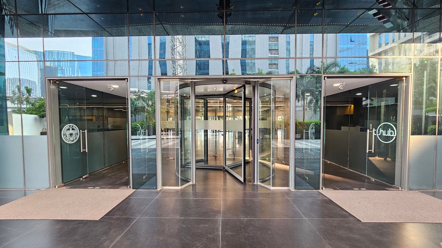 A modern building entrance with a central revolving door flanked by two glass doors. The left door has a logo with text, while the right door features a circular logo with "t-hub". The exterior is reflective glass, showing nearby buildings and greenery. The floor is dark polished stone with beige mats in front of each door. The ceiling is mirrored, reflecting the surroundings.