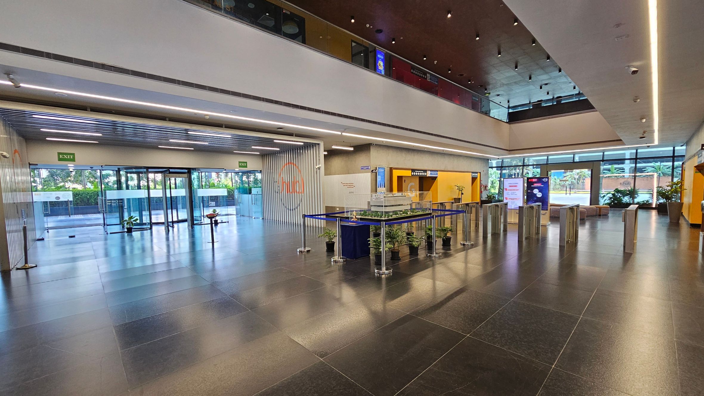A spacious, modern lobby with a dark tiled floor and a high ceiling. On the left, there are glass exit doors with green exit signs above. In the centre, a display area is surrounded by small potted plants and a blue barrier. On the right, there are turnstiles leading to a seating area with large windows showing greenery outside. The ceiling features recessed lighting and a mezzanine level with glass railings.