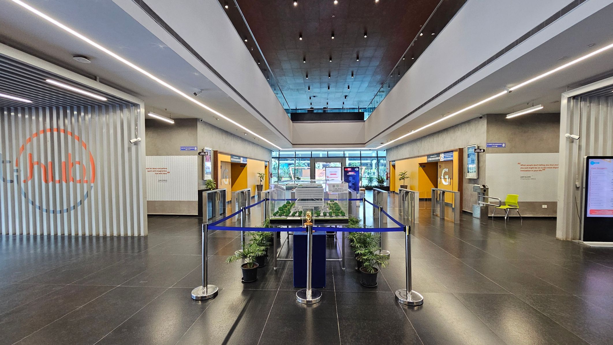 A spacious lobby with a high ceiling and dark tiled floor. In the centre, a model of a building is displayed, surrounded by a blue barrier and potted plants. On the left, a wall with vertical slats and a logo. On the right, a yellow door marked "G" and a green chair. The background features large glass doors and windows, allowing natural light to enter. Overhead, linear lights run along the ceiling.