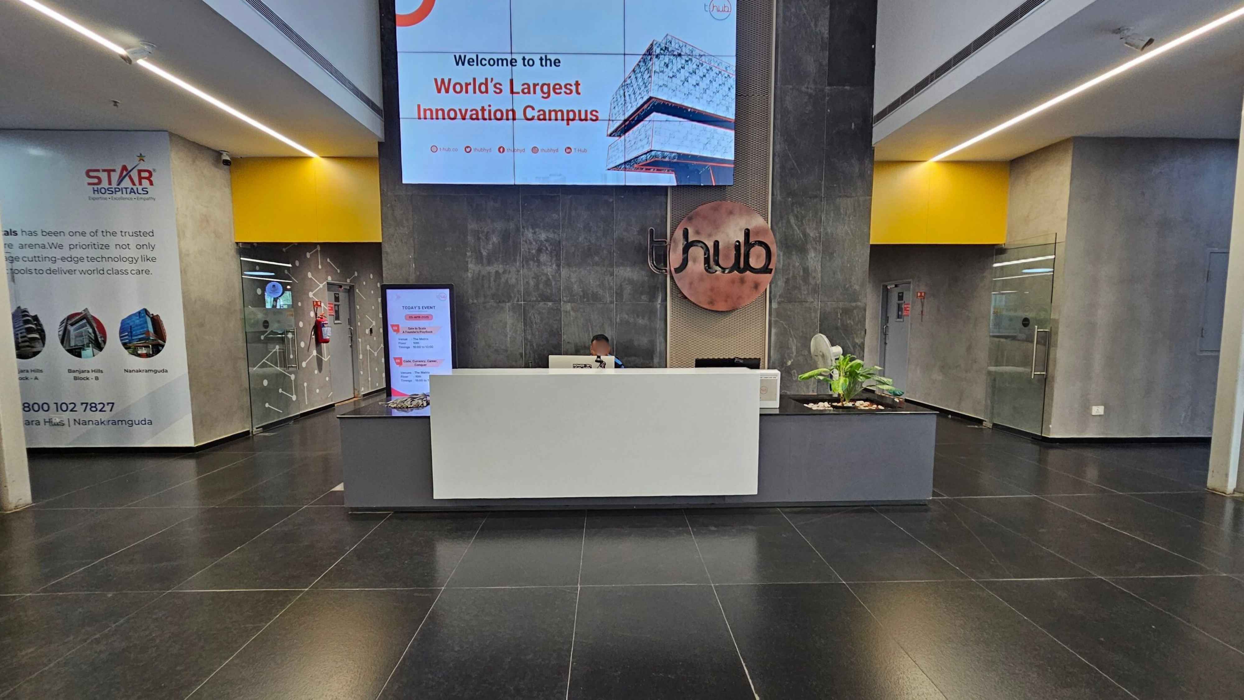 Reception area with a modern design. In the centre, a white desk with a person seated behind it. Above, a large digital screen displays a welcome message. To the right, a glass door and a small plant on the desk. On the left, a wall with promotional material and a digital screen showing event information. The floor is dark and glossy, and the ceiling features linear lighting. The walls are grey with yellow accents.