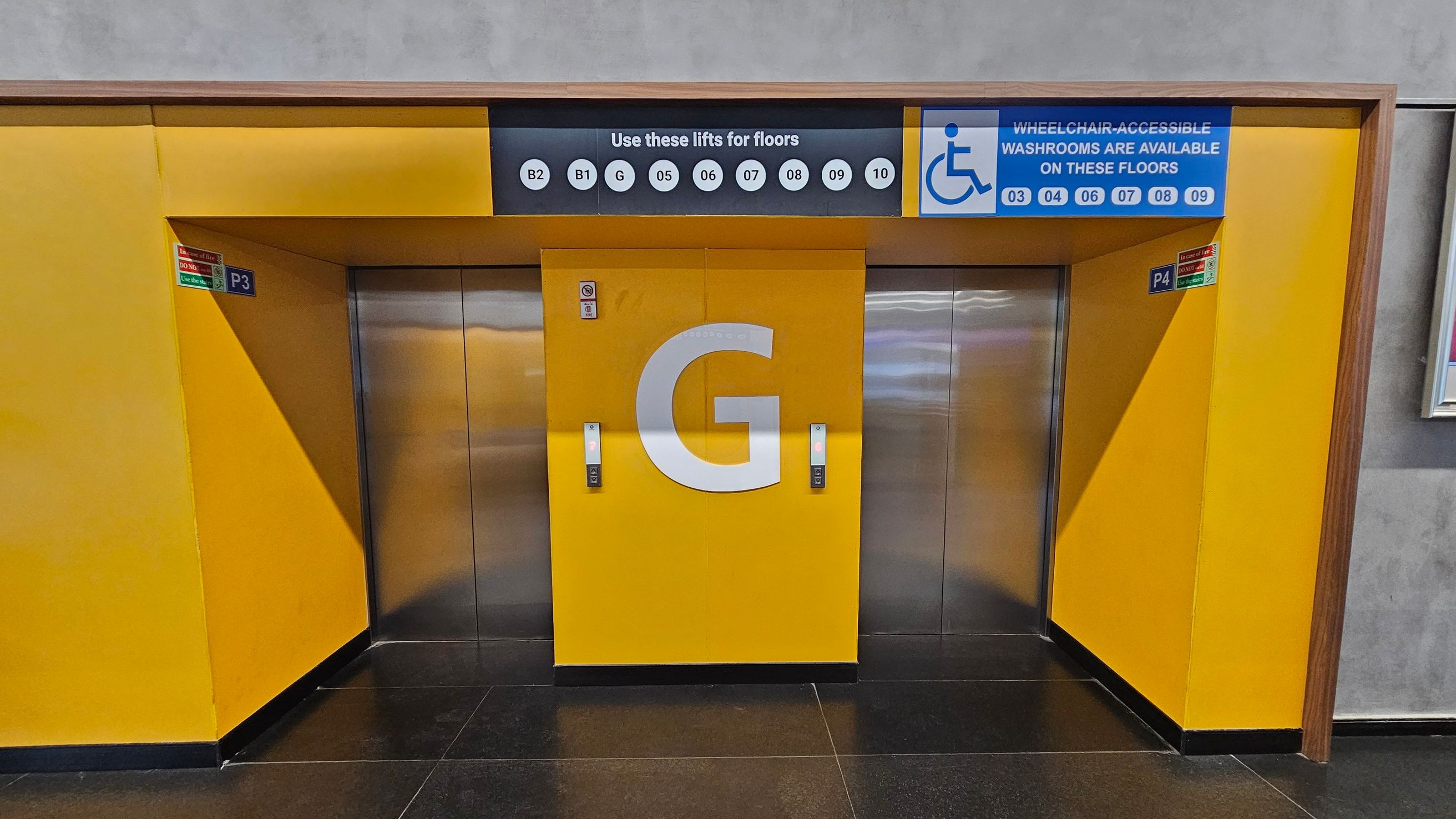 Two lift doors are set in a bright yellow wall. The central lift door features a large letter "G". Above, a sign indicates the floors accessible by these lifts, including B2, B1, G, and floors 05 to 10. A blue sign with a wheelchair symbol indicates wheelchair-accessible washrooms on floors 03, 04, 06, 07, 08, and 09. The floor is dark and glossy, and the ceiling is not visible.