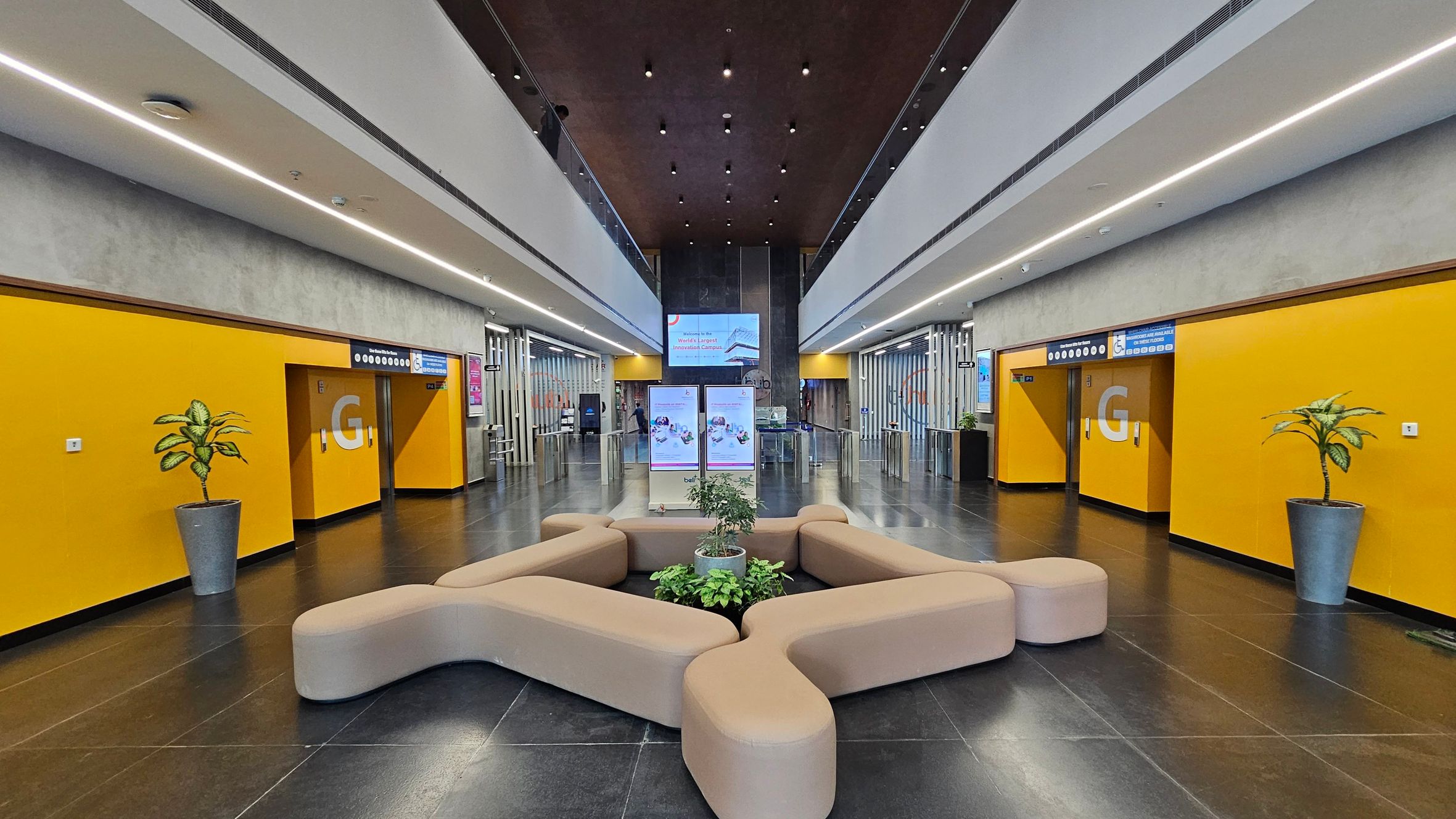 A modern lobby with a symmetrical design. The floor is dark and glossy, and the ceiling features recessed lighting. In the centre, there is a beige, curved seating arrangement surrounding a planter with green plants. The walls on both sides are bright yellow with large "G" letters and potted plants. In the background, there are digital screens and a glass entrance with turnstiles. The upper level has a balcony with glass railings.