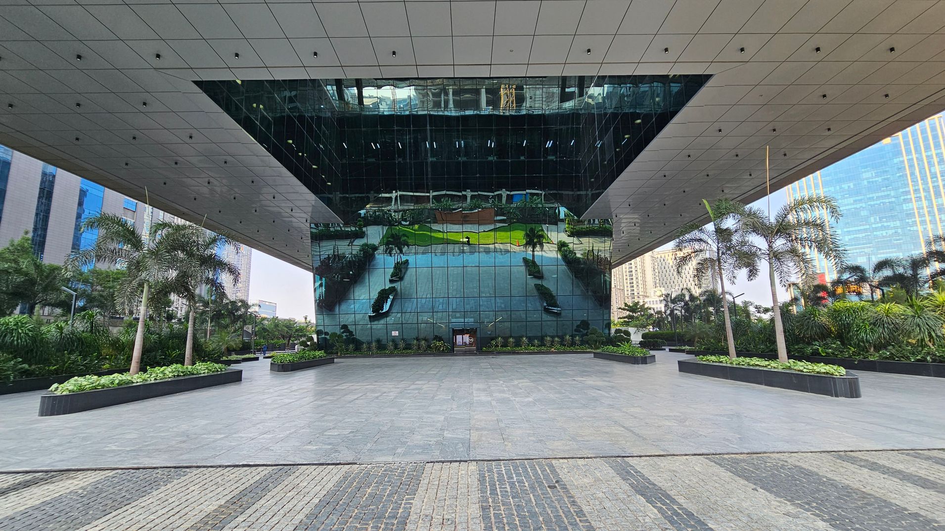 A modern architectural structure with a large reflective glass facade in the centre, mirroring the surrounding area. The foreground features a spacious tiled plaza with a patterned design. On both sides, there are rectangular planters with palm trees and lush greenery. The ceiling is composed of a grid of panels with small lights. In the background, tall buildings are visible, adding to the urban setting.