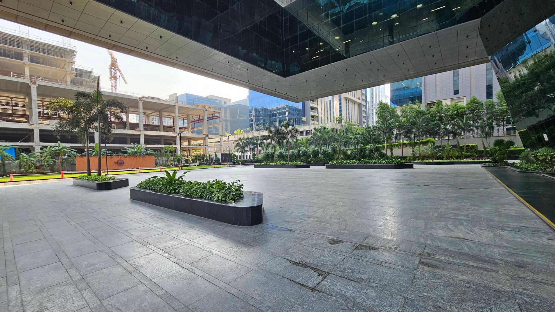 A spacious outdoor area with a tiled floor and a modern architectural ceiling. On the left, there's a construction site with a crane and partially built structures. In the middle, there are rectangular planters with small trees and greenery. On the right, a row of palm trees and other plants lines the edge of the space, with tall glass buildings in the background. The ceiling has recessed lighting and reflective surfaces.
