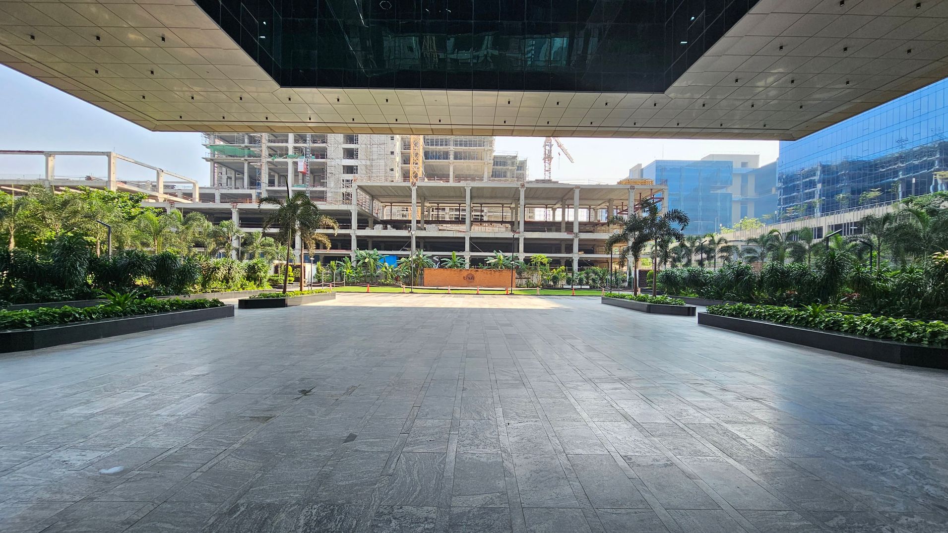 A spacious outdoor area with a tiled floor and a modern ceiling above. In the background, there is a construction site with a partially built structure and a crane. On both sides, there are lush green plants and palm trees lining the walkway. To the right, a glass building reflects the surroundings. The scene is well-lit, suggesting a sunny day.