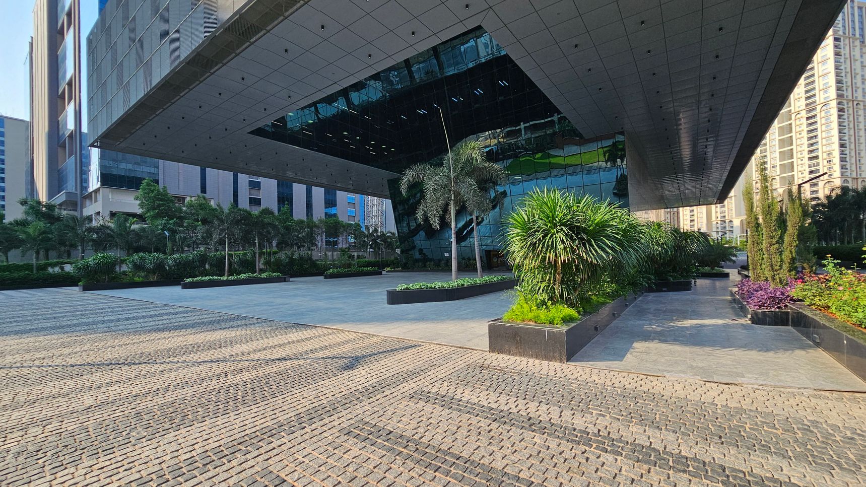A modern architectural structure with a large overhanging roof and reflective glass facade. The foreground features a paved area with a pattern of light and dark stones. On the right, there are landscaped planters with various green plants and shrubs. The background includes tall buildings and more greenery, with trees lining the left side. The ceiling of the overhang is composed of dark panels with small lights.