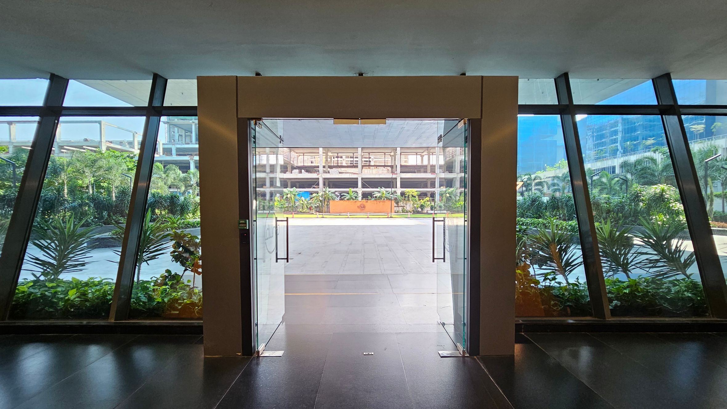 A glass door in the centre opens to an outdoor area with a tiled floor. On either side, large glass panels provide a view of lush green plants and trees. The background features a modern building with multiple floors and a clear, structured design. The floor inside is dark and glossy, while the ceiling is plain and light-coloured.