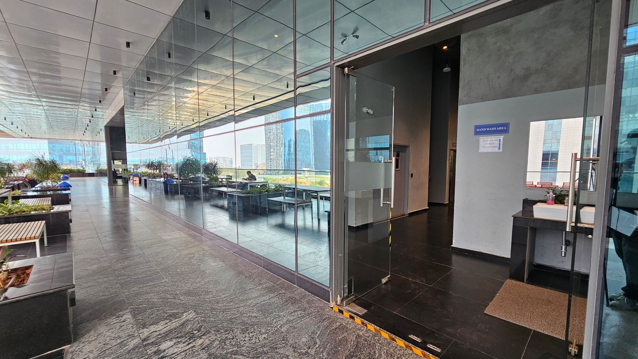 A modern indoor space with a reflective glass wall on the left, showing a corridor lined with potted plants and seating areas. The floor is made of dark stone tiles. On the right, there's an open door leading to a hand wash area with a sink and a sign above it. The ceiling is sleek and metallic, with recessed lighting. In the background, large windows reveal a view of tall buildings outside.