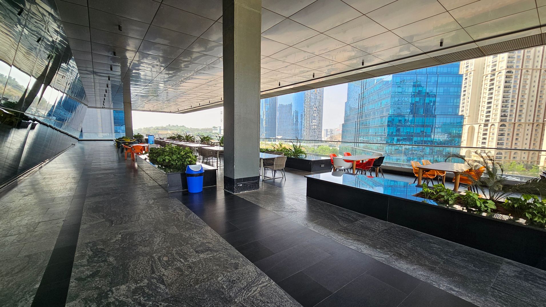 A modern outdoor terrace with a sleek, reflective ceiling and dark stone flooring. On the left, a mirrored wall reflects the surroundings. In the middle, there are tables and chairs, some with orange and red seats, surrounded by planters with green foliage. A blue bin is visible near the centre. The right side features a glass railing overlooking a cityscape with tall, blue-glass buildings and a beige high-rise.