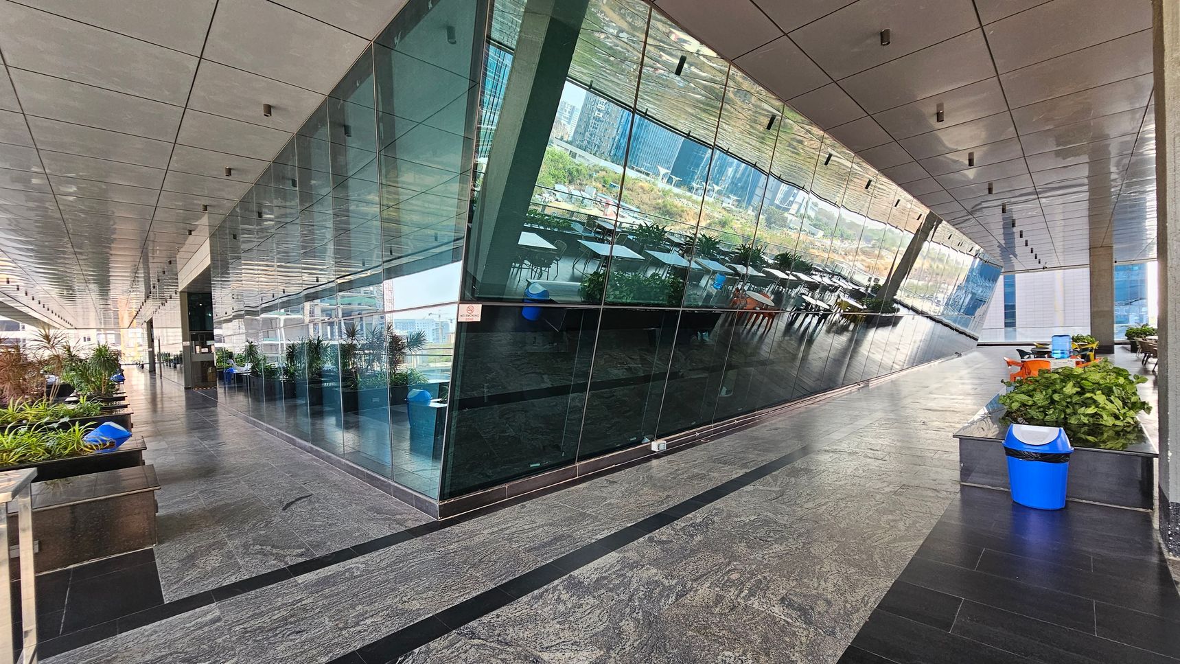 A modern indoor space with a reflective glass wall on the right, showing outdoor scenery. The left side features a row of potted plants and blue bins along a grey stone floor. The ceiling is lined with recessed lights. In the background, there are tables and chairs, suggesting a seating area. The overall design is sleek and contemporary.