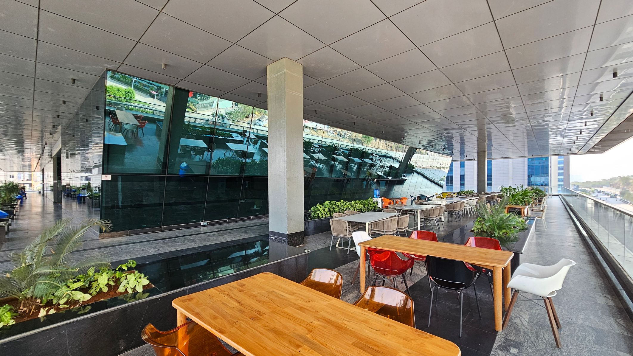 A modern indoor space with a reflective glass wall on the left, showing outdoor scenery. In the centre, there are wooden tables with a mix of red, black, and white chairs. The right side features a glass railing overlooking a cityscape. The floor is dark grey, and the ceiling is covered with light-coloured panels. Green plants are placed along the edges, adding a touch of nature to the sleek, contemporary design.