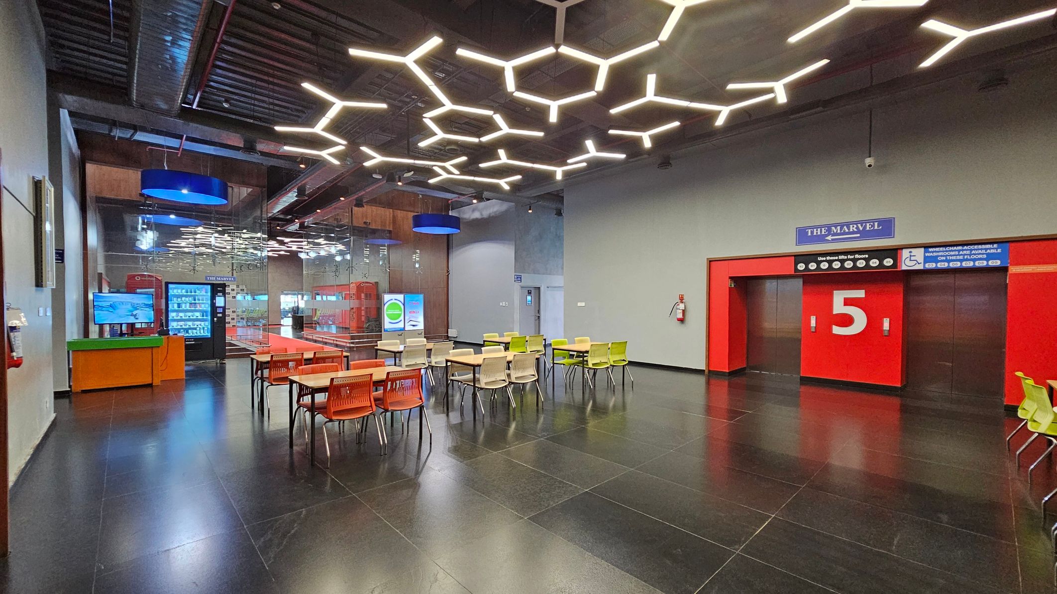 A modern interior with a dark tiled floor and a ceiling featuring geometric light fixtures. On the right, there are red doors marked with the number 5 and a sign above reading "The Marvel". In the centre, tables and chairs in orange and green are arranged. On the left, a vending machine and a screen are visible near a glass wall. Blue circular lights hang from the ceiling, and a fire extinguisher is mounted on the wall.