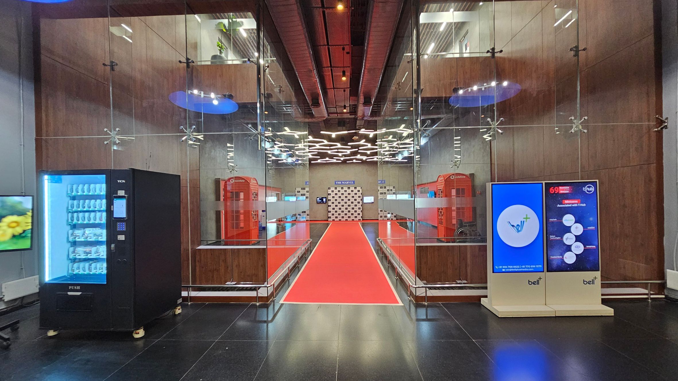 A modern interior with a black vending machine on the left and a digital display on the right. The floor is black with a red carpet running down the centre. The walls are glass and wood, with two red telephone booths visible in the background. The ceiling features geometric lighting fixtures. A sign at the end of the carpet reads "THE MARVEL" with a checkered backdrop. The space has a sleek, contemporary design with a mix of technology and traditional elements.