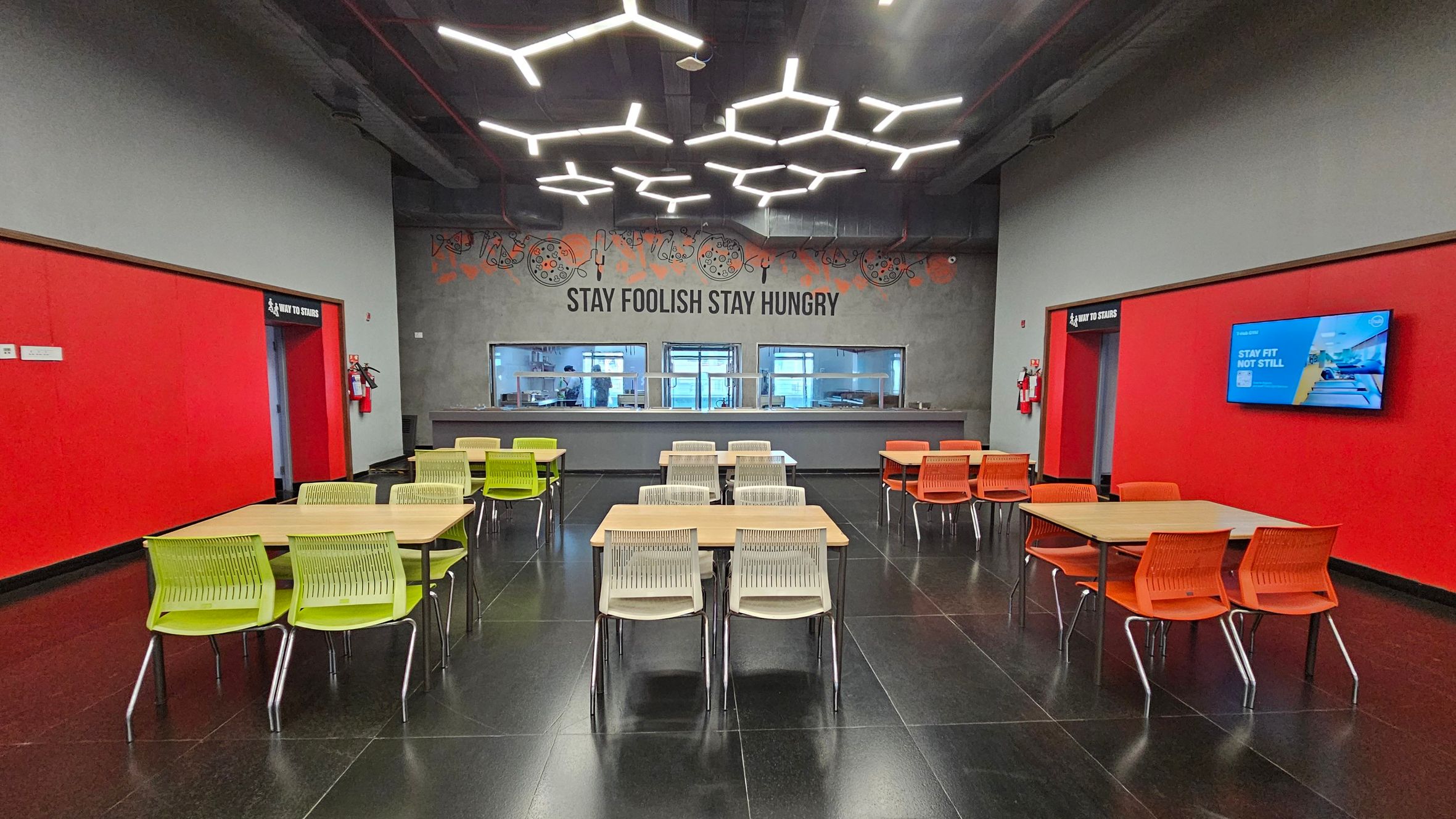 A modern cafeteria with a dark floor and a grey ceiling featuring geometric light fixtures. In the centre, there are tables with green and orange chairs. The back wall is grey with a motivational quote and windows to a kitchen area. The left and right walls are red, with a doorway on each side. The right wall also has a digital screen displaying a message.