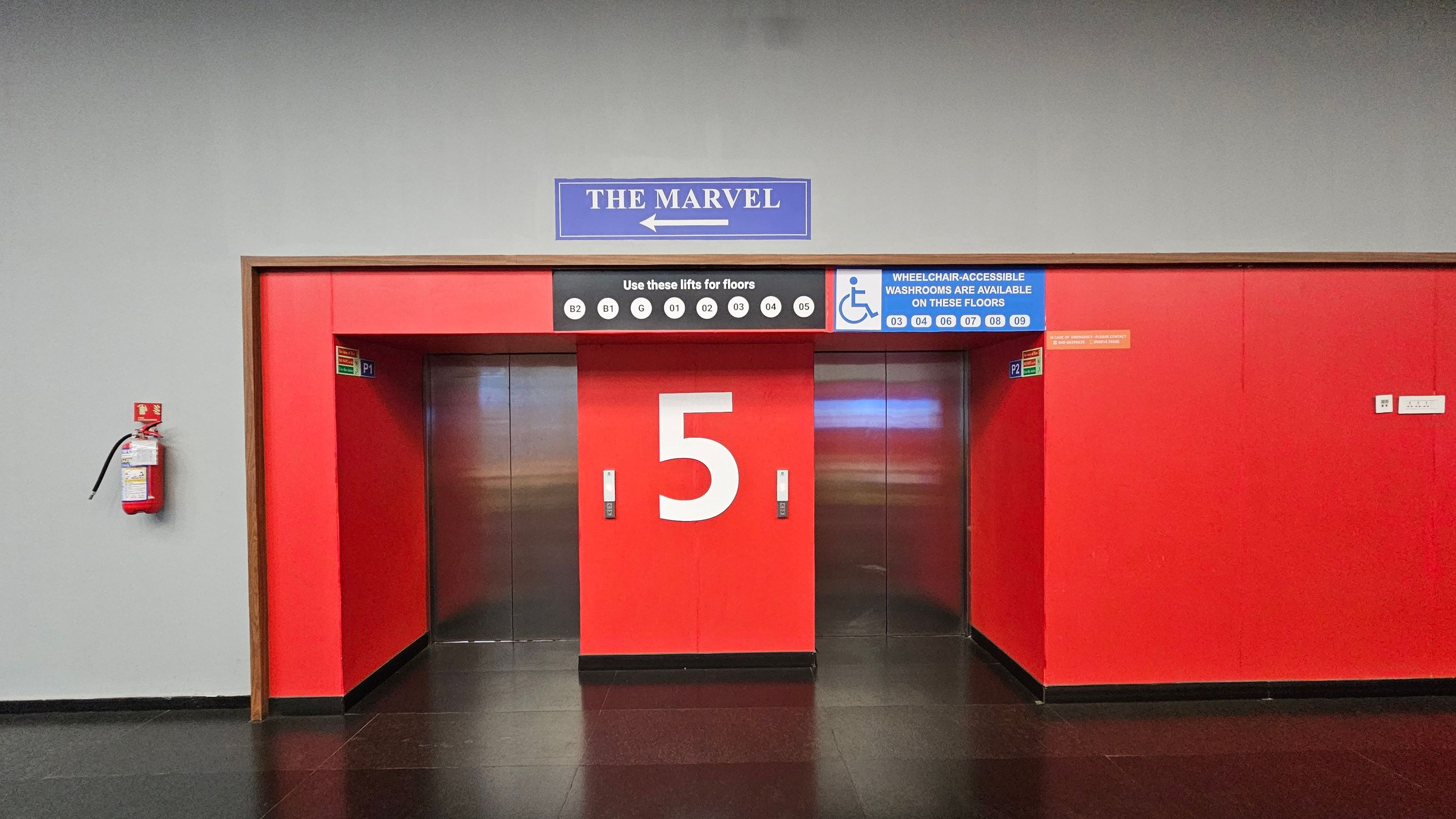 A set of three lifts with red doors, the central one prominently displaying the number "5". Above, a sign reads "THE MARVEL" with an arrow pointing left. Another sign indicates floors accessible by these lifts, including wheelchair-accessible washrooms on certain floors. A fire extinguisher is mounted on the left wall. The floor is dark and shiny, and the walls are grey and red.