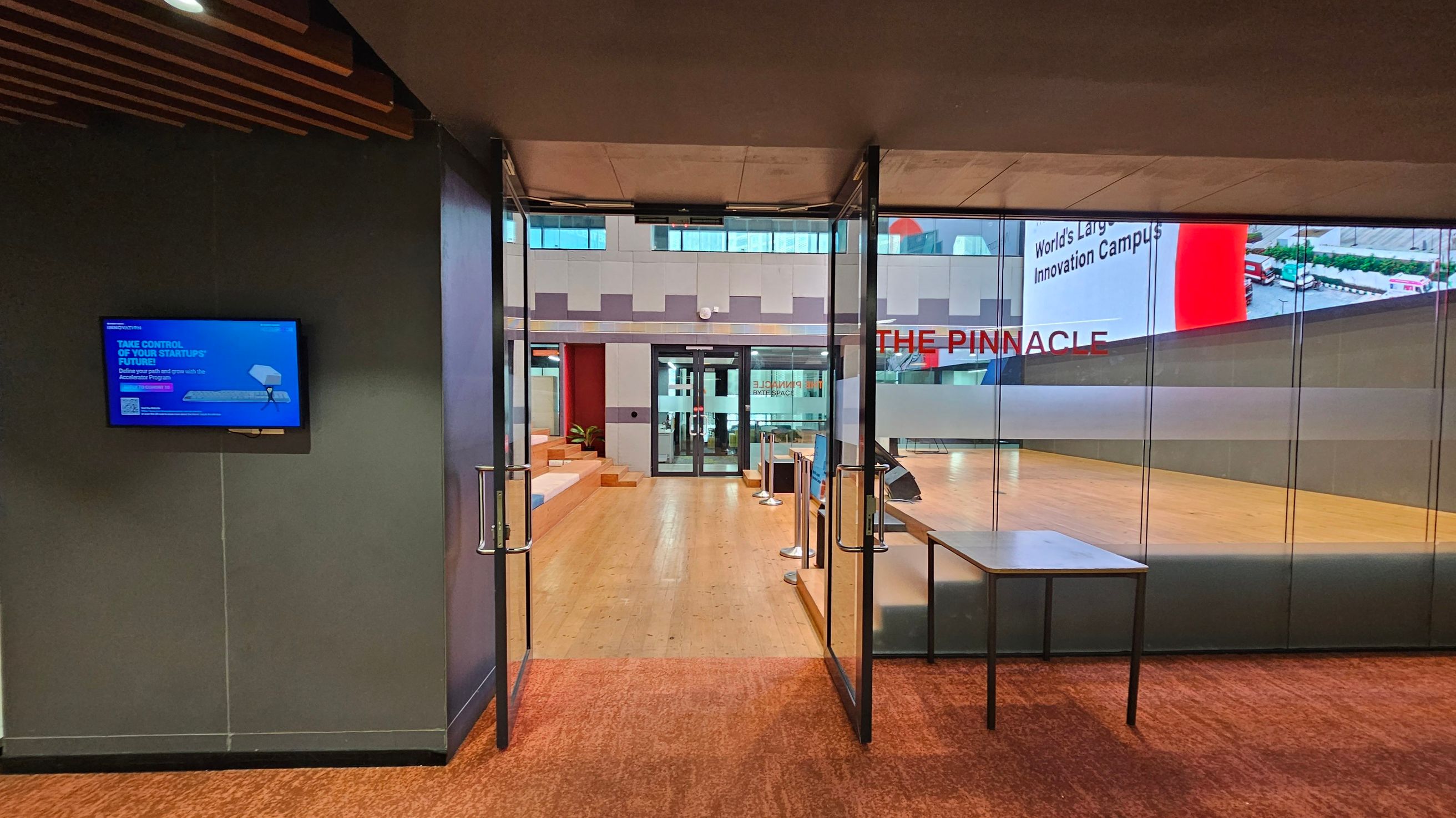 A modern interior space with a wooden slatted ceiling and a mix of orange and grey flooring. On the left, there are two green chairs, a fire extinguisher, and a digital display stand. A small plant is placed on a shelf above. In the centre, a glass door leads to another room. On the right, a water cooler and a bin are against a grey wall. The lighting is bright, with several linear lights on the ceiling.