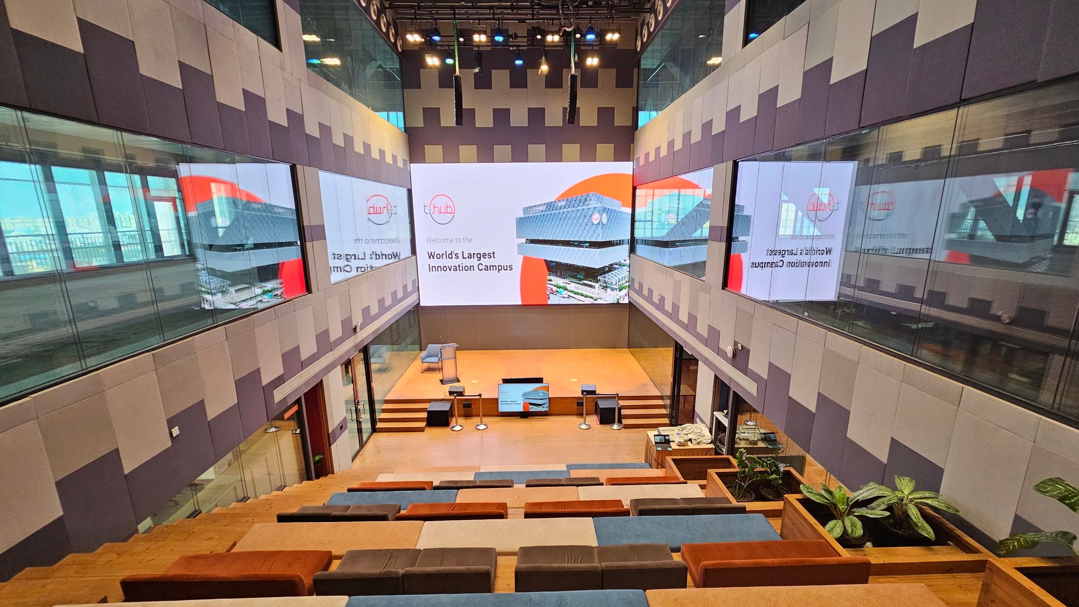 A modern auditorium with tiered seating featuring multicoloured cushions. The seating area descends towards a stage with a podium and a large screen displaying a building and text about an innovation campus. The walls are lined with reflective glass panels, and there are potted plants near the seating. The ceiling has spotlights and speakers, and the floor is wooden.
