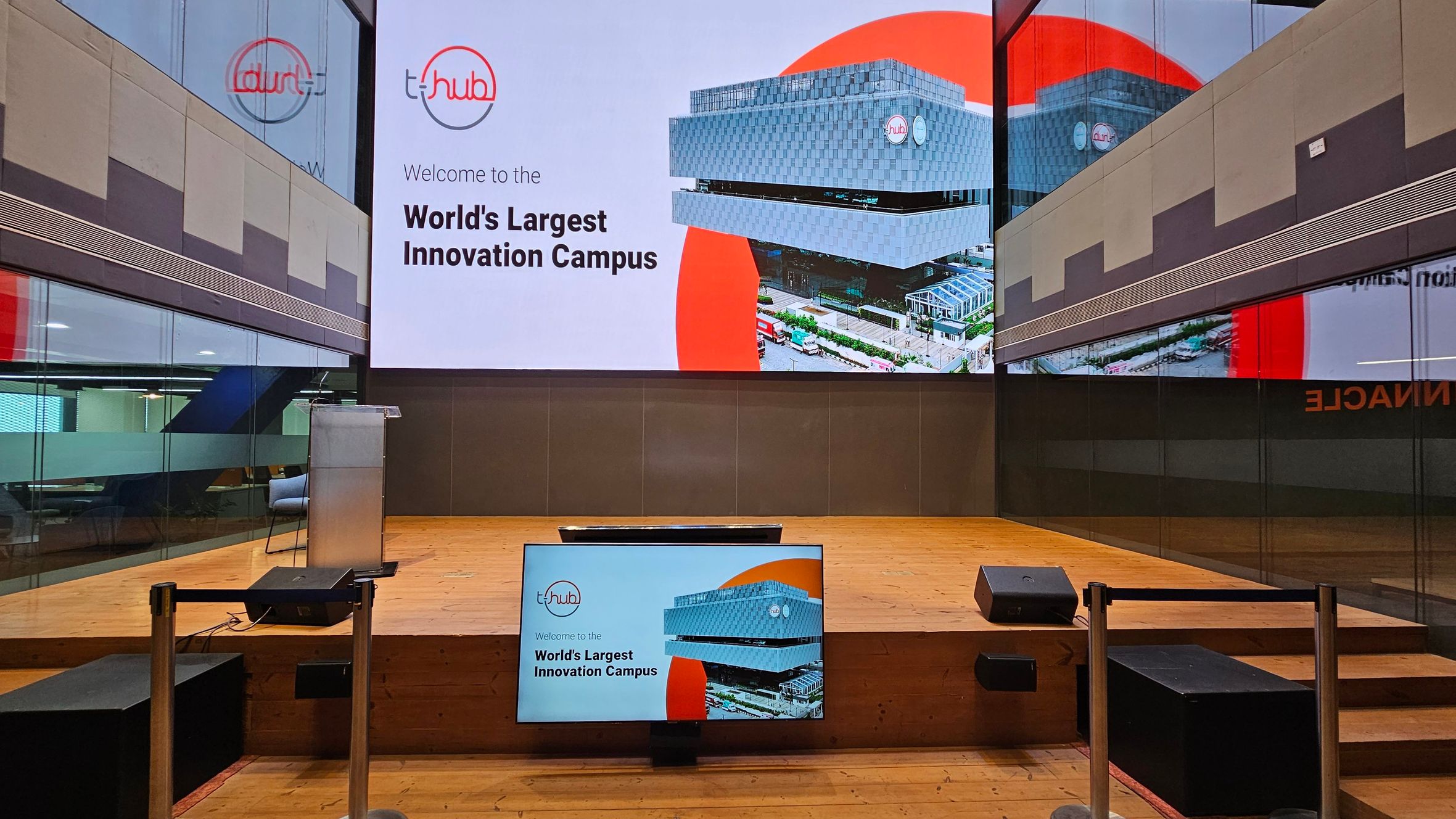 A modern indoor auditorium with a wooden stage. At the centre, a large screen displays "Welcome to the World's Largest Innovation Campus" with an image of a building. Below, a smaller screen mirrors the same content. A podium is positioned on the left side of the stage. The walls are made of reflective glass panels, showing partial reflections of the interior. The floor is wooden, and there are speakers on either side of the stage.