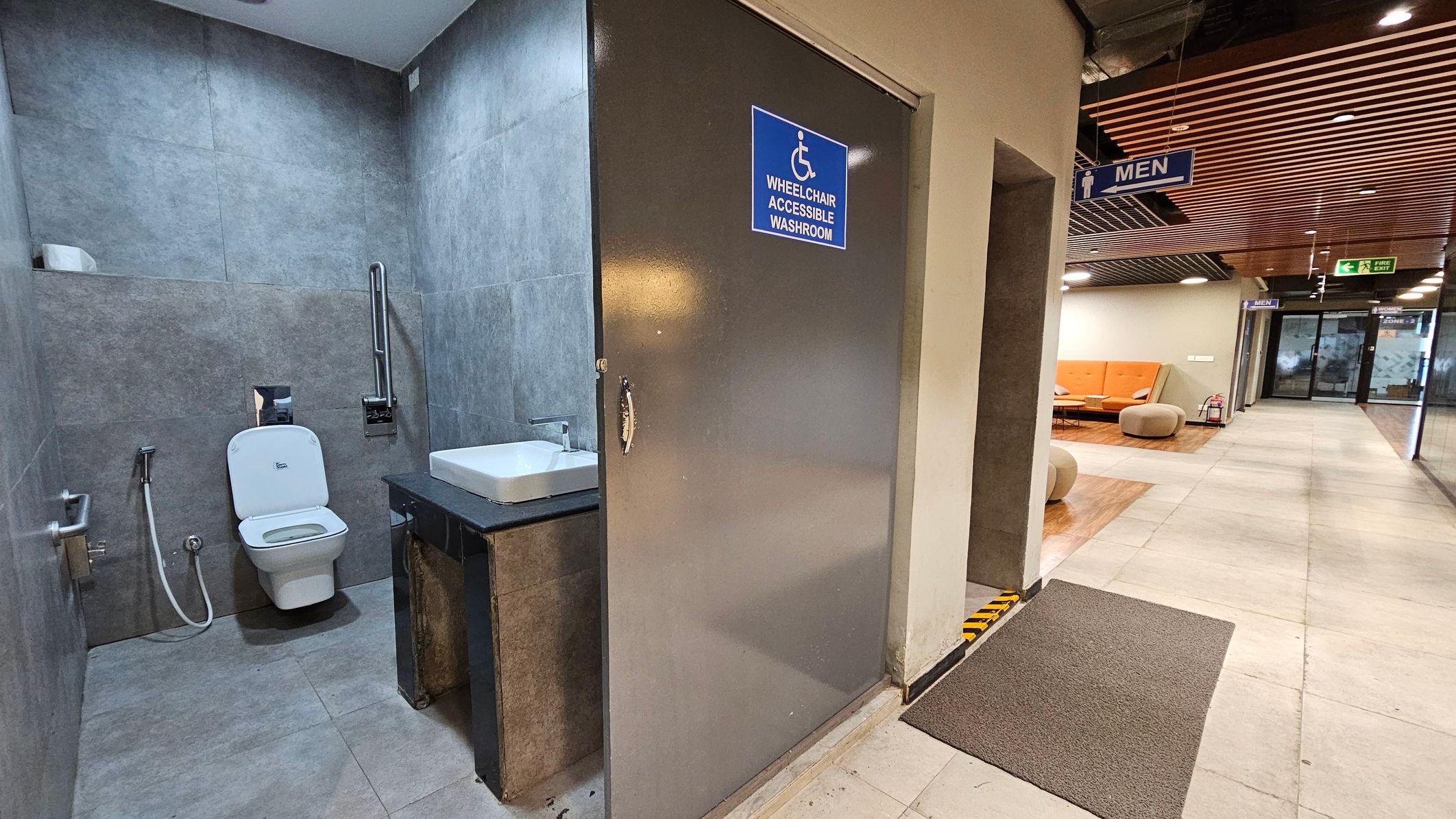 A wheelchair-accessible washroom with a grey door and a blue sign is on the left. Inside, there's a white toilet with a grab bar and a sink with a black countertop. The floor is tiled in grey. To the right, a corridor leads to a seating area with orange sofas and round beige stools. The ceiling has wooden slats, and there's a "Men" sign hanging above. The floor is light-coloured tile with a grey mat near the washroom entrance.
