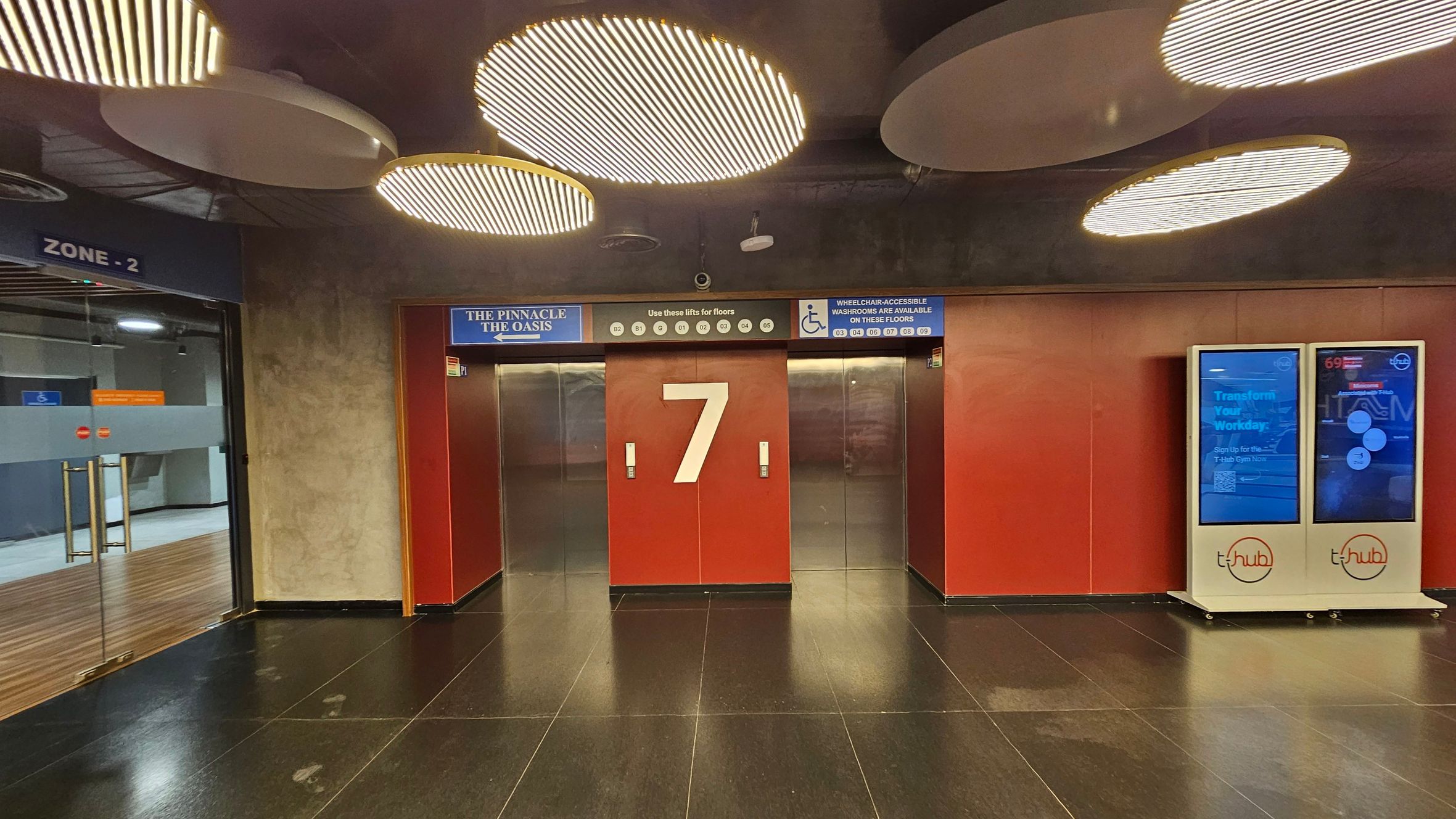A modern interior with two metallic lift doors in the centre, marked by a large number "7" on a red panel. Above, signs indicate lift access to various floors and wheelchair-accessible washrooms. To the right, two digital screens display information, branded with "t-hub". On the left, a glass door leads to "Zone 2", with a wooden floor visible beyond. The ceiling features circular light fixtures, and the floor is dark and glossy.