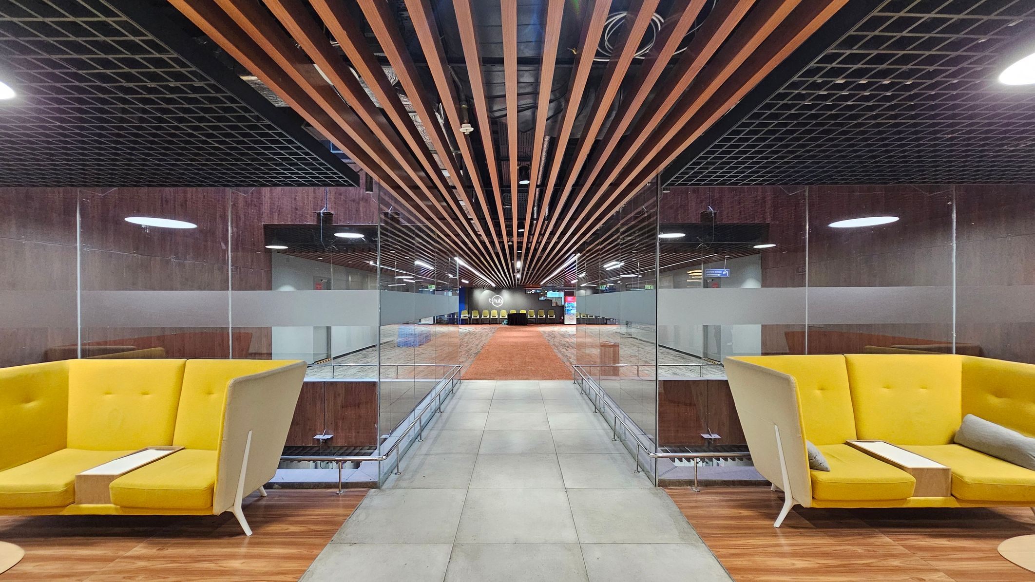 A modern interior with a symmetrical design. On the left and right, there are yellow, high-backed sofas with small tables attached. The floor is a mix of grey tiles and wood. The ceiling features a grid pattern with wooden slats running lengthwise. In the centre, a glass partition separates the seating area from a spacious hallway leading to a distant wall with a logo and a row of chairs. The lighting is bright, with circular fixtures on the ceiling.