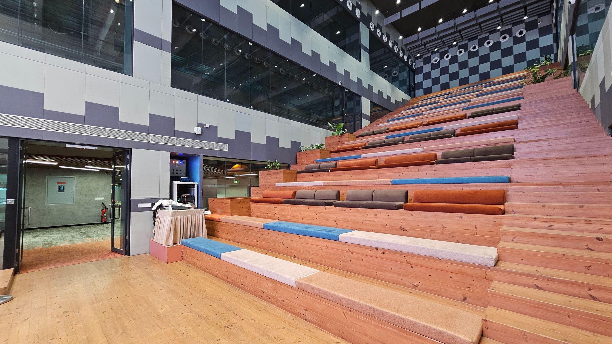 A modern interior space with a large, stepped seating area on the right, featuring wooden benches with colourful cushions in shades of blue, orange, and brown. On the left, there is a glass entrance with a door leading to another room. Above the entrance, the wall is decorated with a geometric pattern in grey tones. The ceiling is high with visible lighting fixtures and a checkered pattern on the upper wall. The floor is wooden, matching the seating area.