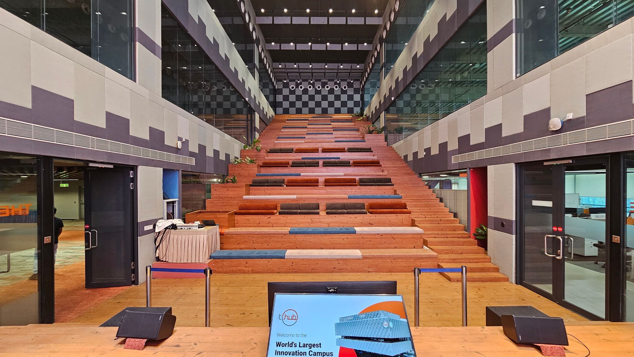 A large, open interior space with a wide wooden staircase in the centre, featuring colourful cushions. The walls on either side are grey with a geometric pattern, and glass panels line the upper sections. The ceiling is high with visible lighting fixtures. On the floor, there is a digital display screen in the foreground, and a small table with equipment to the left. Doors on both sides lead to adjacent rooms, and there are some potted plants along the stairs.