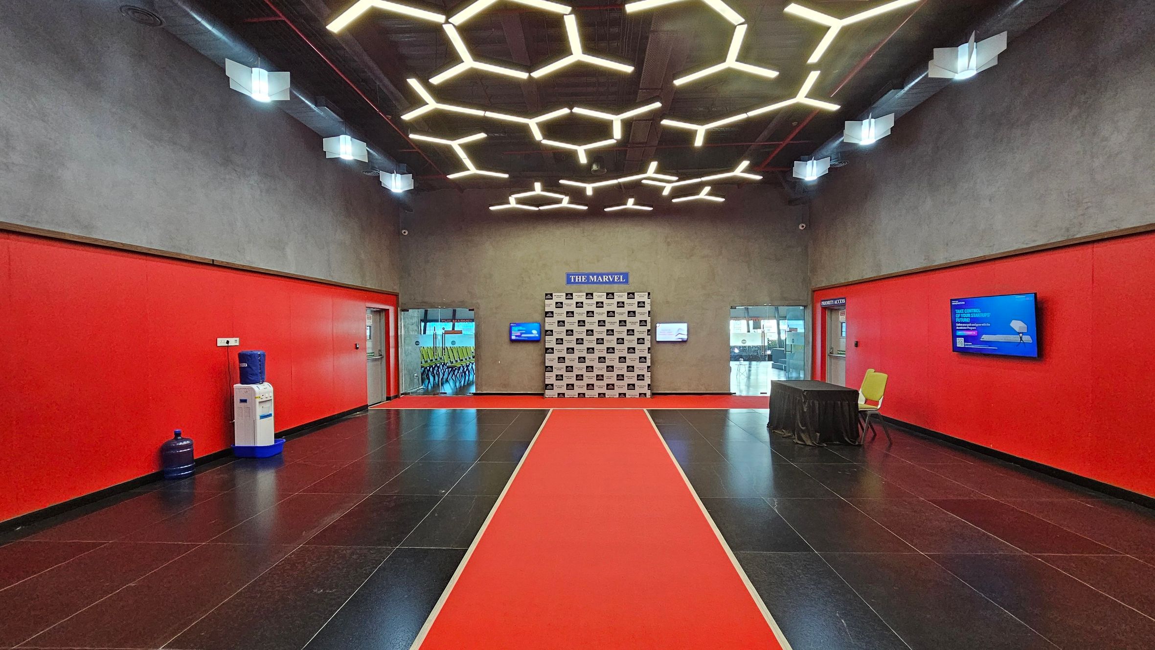A spacious room with red walls and a black tiled floor. In the centre, a red carpet leads to a backdrop labelled "The Marvel". The ceiling features hexagonal light fixtures. On the left, a water cooler and a large water bottle are against the wall. On the right, a table with a black cloth and a yellow chair are near a wall-mounted screen. Two glass doors on either side of the backdrop reveal a room with yellow chairs.