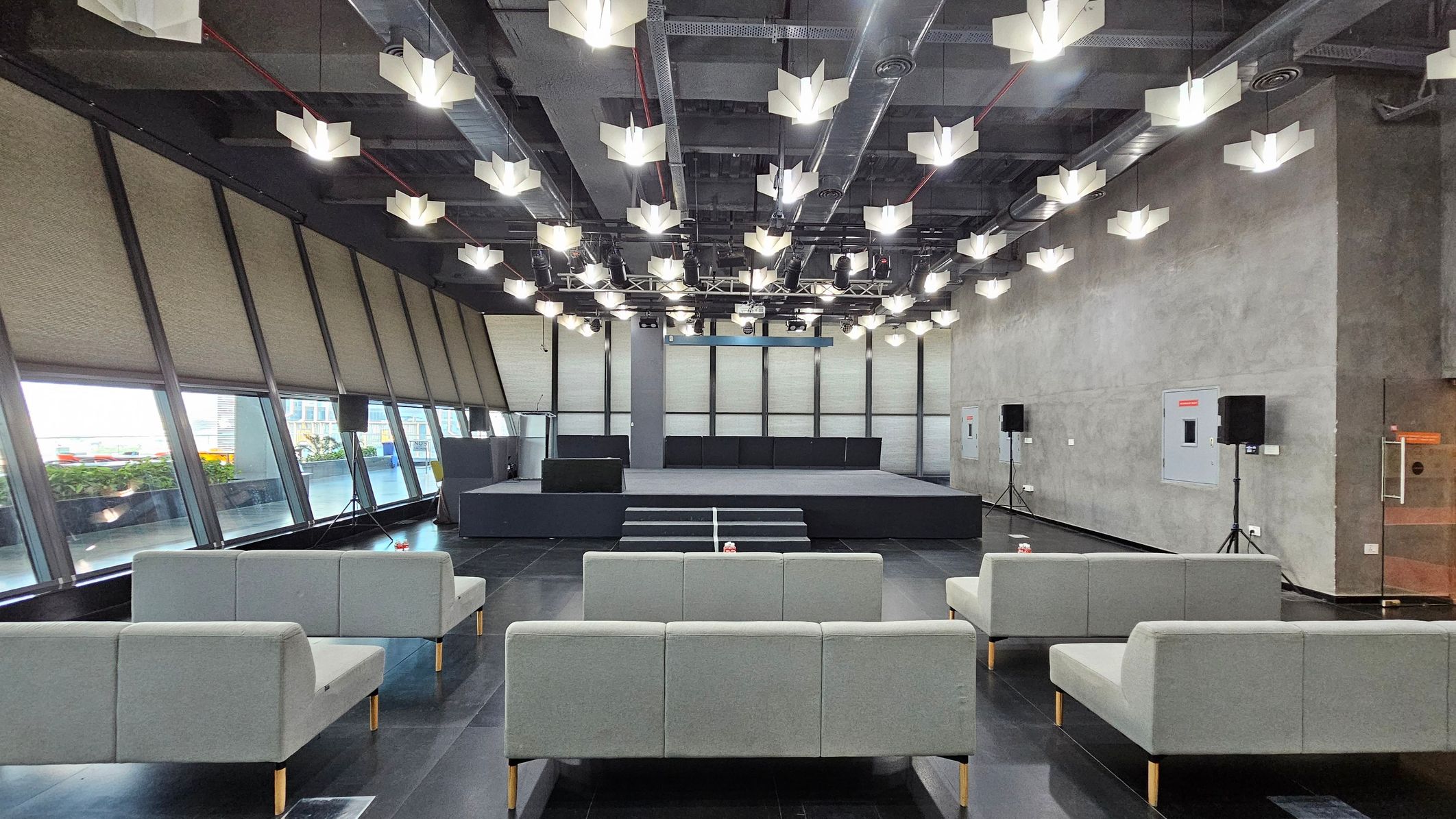 A modern interior space with a stage at the centre, featuring a set of steps leading up to it. The ceiling is adorned with geometric light fixtures. On the left, large slanted windows with blinds allow natural light in, while the right wall is plain with mounted speakers. The floor is dark and glossy, with several grey sofas arranged in rows facing the stage.