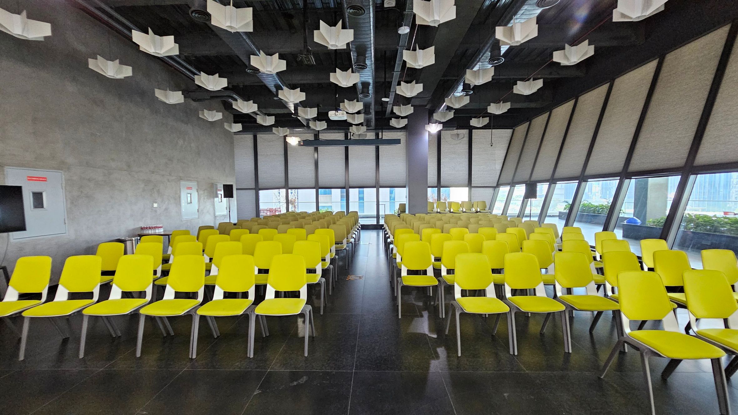 A room with rows of yellow chairs arranged in a grid pattern on a dark tiled floor. The ceiling is adorned with white, angular light fixtures. The left wall is plain with a few panels, while the right side features large, slanted windows with blinds, allowing some natural light to enter. At the back, more chairs are visible, and outside the windows, there are glimpses of a building and some greenery.
