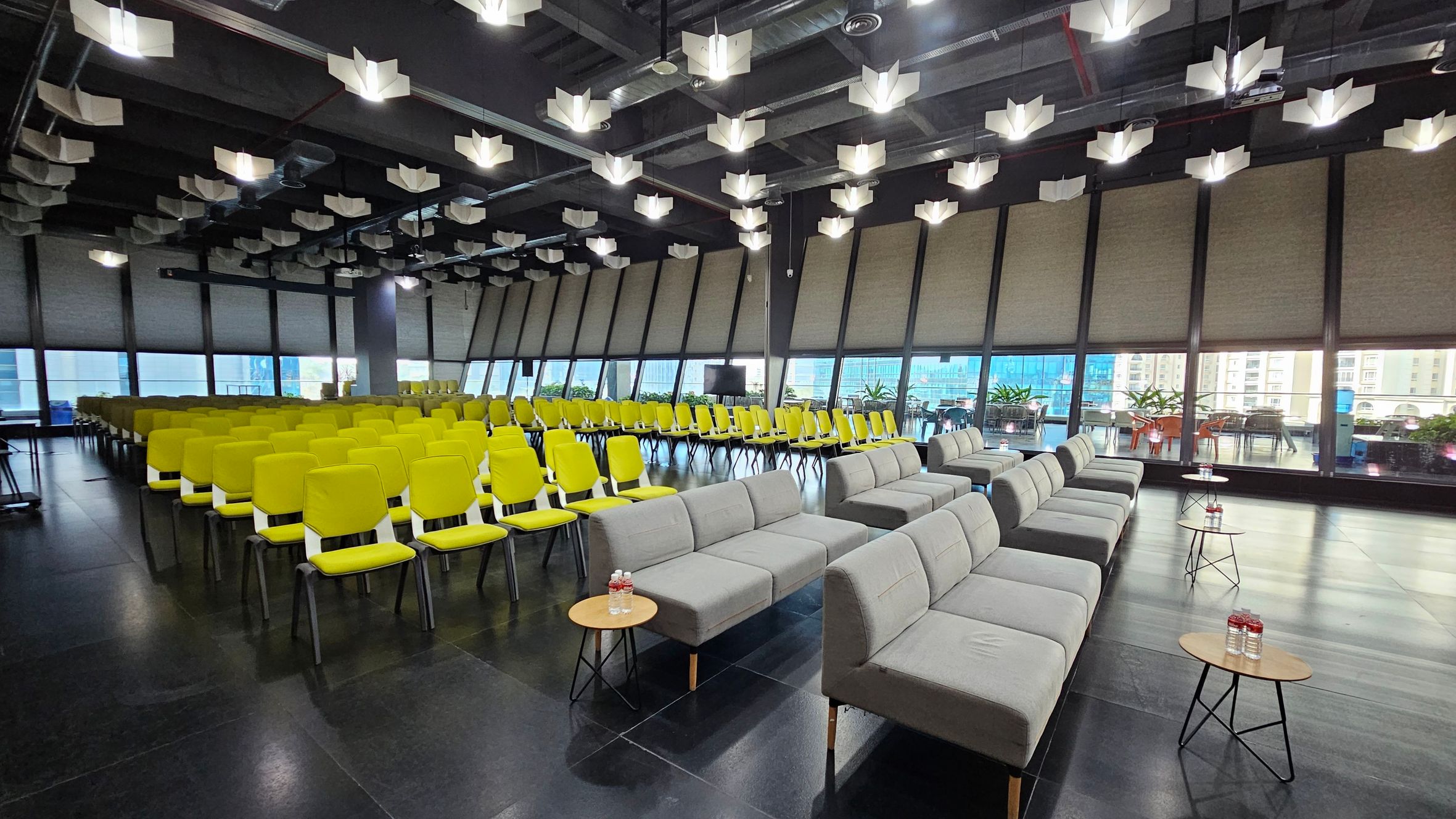 A spacious conference room with rows of bright yellow chairs on the left and grey sofas on the right. Small round tables with bottled water are placed in front of the sofas. Large windows in the background reveal a view of buildings and outdoor seating with plants. The ceiling features modern, geometric light fixtures, and the floor is dark and polished.