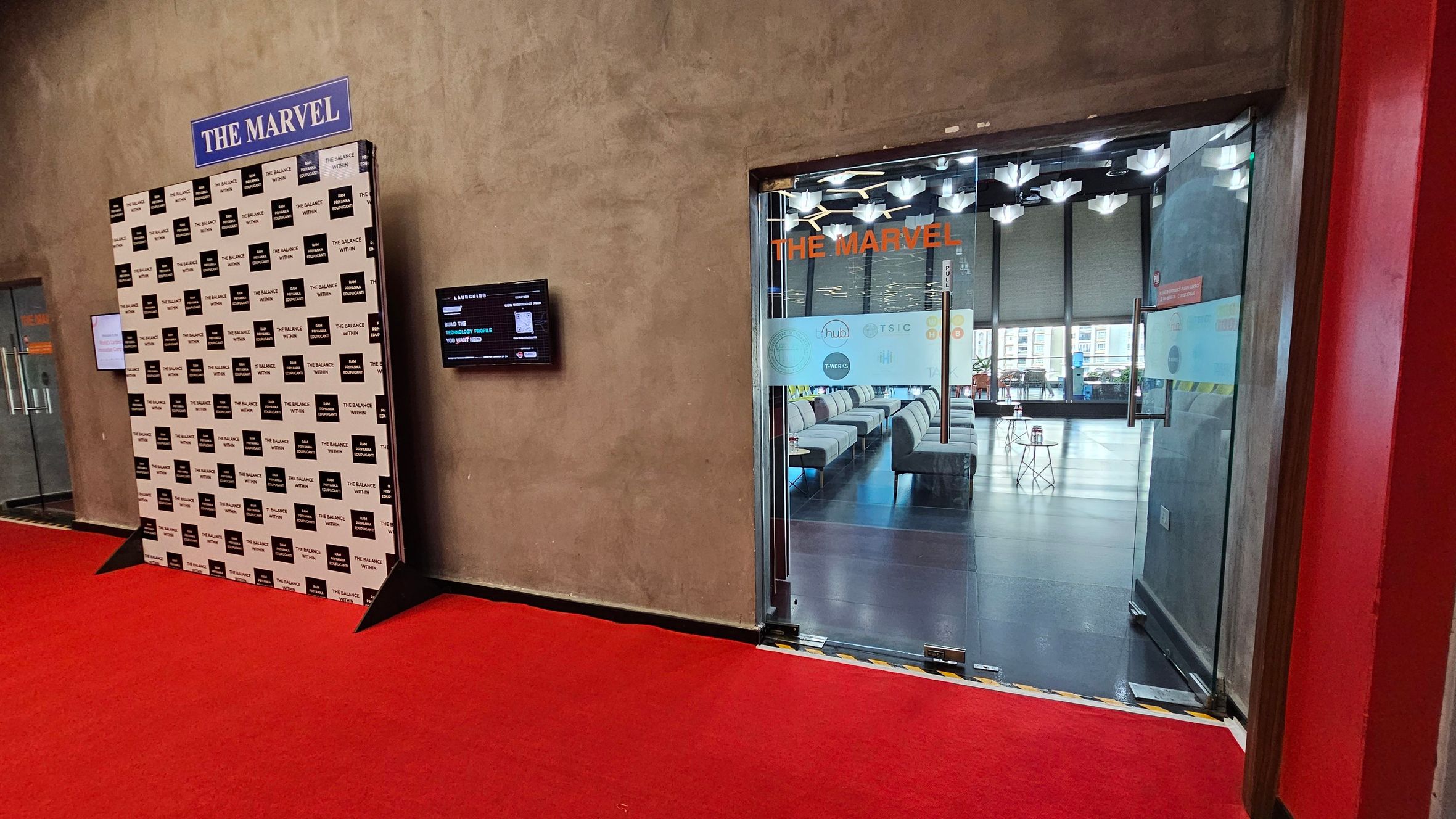 A hallway with a red carpeted floor. On the left, a backdrop with "THE BALANCE NETWORK" repeated in a pattern. A digital screen is mounted on the wall next to it. On the right, a glass door with "THE MARVEL" in orange letters leads into a room with modern seating and geometric ceiling lights. The room has large windows in the background.