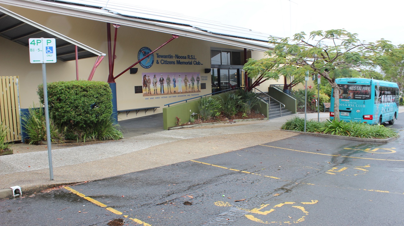 The front wall with military artwork, the Tewantin Noosa RSL badge, two etched tiles with inscriptions, and blue-painted seats on the yellow wall under a facade. The front door is accessed through a two-side ramp access walkway and a stepped walkway in the centre flanked by green-painted half-walls and steel railings facing a sidewalk and a parking lot. The wall is surrounded by sculpted areas of grass and a tree. Three direction signs are visible. The front wall with military artwork, the Tewantin Noosa RSL badge, two etched tiles with inscriptions, and blue-painted seats on the yellow wall under a facade. The front door is accessed through a two-side ramp access walkway and a stepped walkway in the centre flanked by green-painted half-walls and steel railings facing a sidewalk and a parking lot. The wall is surrounded by sculpted areas of grass and a tree. Three direction signs are visible.