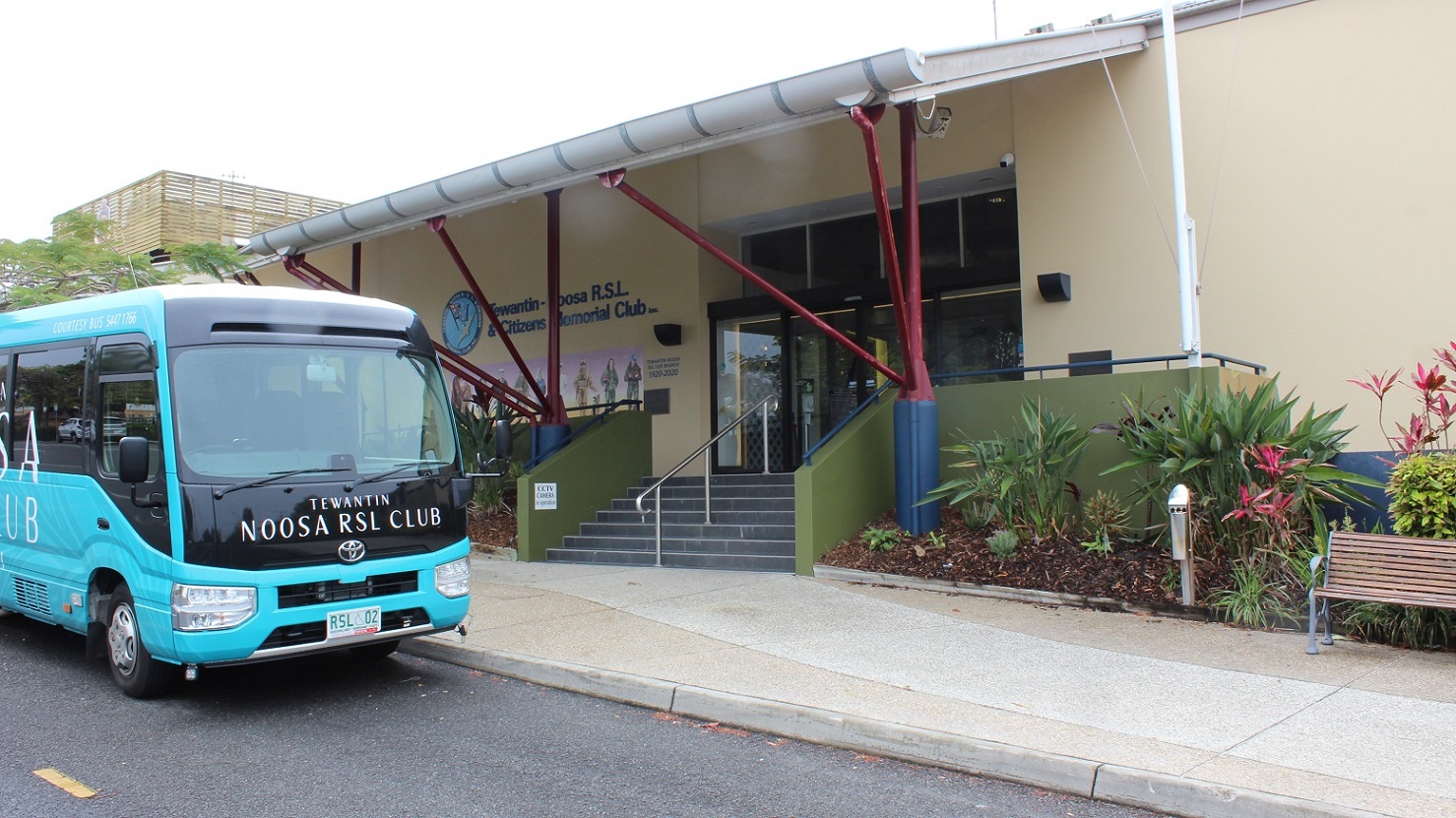 Another view of the previous image focuses on the glass sliding door and black tiled steps under the red/blue pillar-supported exterior. A steel rail in the centre divides the stepway into two halves bordered by green-painted half-walls with rails on both sides. Plants are in triangle forms by a seat on the right facing the sidewalk and parking lot. The bus is packed in front. Another view of the previous image focuses on the glass sliding door and black tiled steps under the red/blue pillar-supported exterior. A steel rail in the centre divides the stepway into two halves bordered by green-painted half-walls with rails on both sides. Plants are in triangle forms by a seat on the right facing the sidewalk and parking lot. The bus is packed in front.
