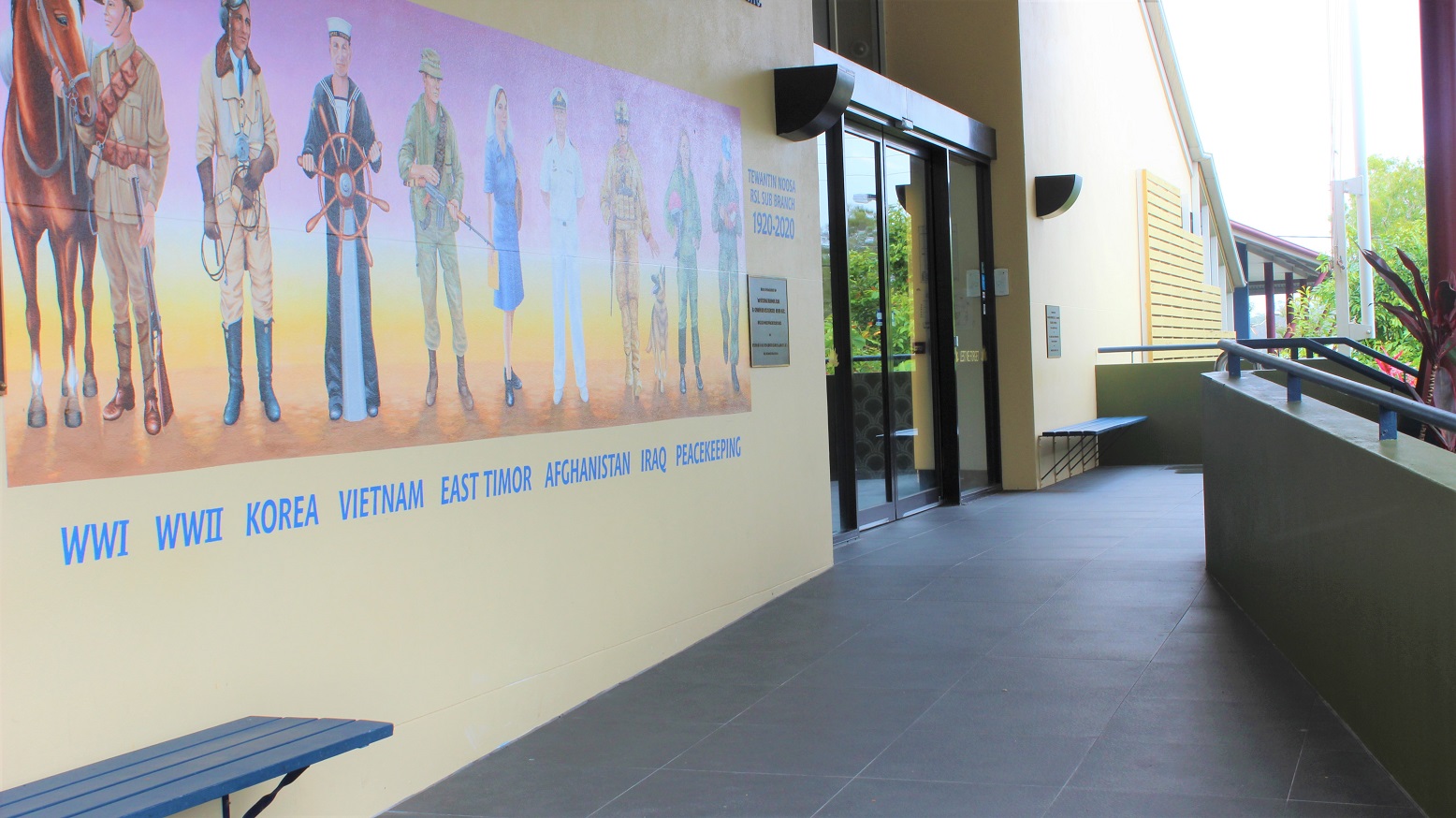 A closer view of the yellow wall with military artwork, engraved inscriptions, and blue wall seats on both sides on a grey-tiled walkway flanked by green-painted half-walls with railings. A closer view of the yellow wall with military artwork, engraved inscriptions, and blue wall seats on both sides on a grey-tiled walkway flanked by green-painted half-walls with railings.