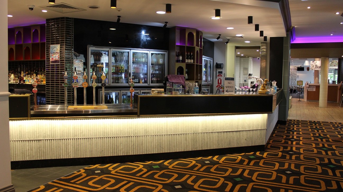 A view of the cafe's counter, designed in black and white and has two sets of broad fridges packed with drinks separated by a wine rack behind the counter. On the left by a pillar is another wine rack packed with wines. On the counter are a screen, infographic, pos, sanitizers, and other items on the right. Beers on tap steel covers a lower spot on the counter. The floor is covered in an army theme. The walkway leads to the bistro-themed restaurant. A view of the cafe's counter, designed in black and white and has two sets of broad fridges packed with drinks separated by a wine rack behind the counter. On the left by a pillar is another wine rack packed with wines. On the counter are a screen, infographic, pos, sanitizers, and other items on the right. Beers on tap steel covers a lower spot on the counter. The floor is covered in an army theme. The walkway leads to the bistro-themed restaurant.