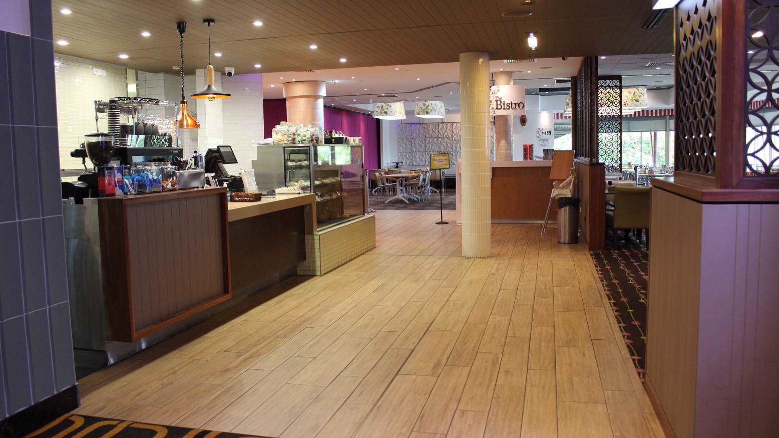 The Bistro restaurant is accessed through a beige-covered floor, with a brown designed counter on the left having coffee packs,a cabinet with cups, a checking screen, and a lit snacks showing cabinet. On the left is a wide entrance with walled space having a rubbish bin and seats. Two white pillars face the bistro tag on a flowering white lamp cover and the direction board on the entry to the purple and white wall dining room with seats and tables on the right. The Bistro restaurant is accessed through a beige-covered floor, with a brown designed counter on the left having coffee packs,a cabinet with cups, a checking screen, and a lit snacks showing cabinet. On the left is a wide entrance with walled space having a rubbish bin and seats. Two white pillars face the bistro tag on a flowering white lamp cover and the direction board on the entry to the purple and white wall dining room with seats and tables on the right.
