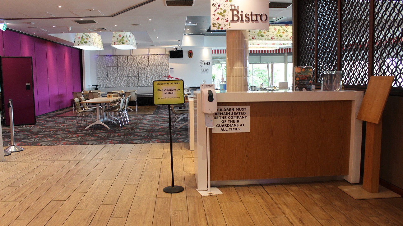 A close-up view of the Bistro restaurant. On the right, the Bistro is written on a lamp cover by a pillar on a beige-tiled floor with a brown wooden accented counter. On the desk is an infographic, a menu, and hand sanitizer. Direction board, a sanitizer dispenser, and a wooden board are seen by the counter. The hallway leads to a purple and white marble wall by the seating area with sets of four brown tables arranged around a table on grey and red tiles. A close-up view of the Bistro restaurant. On the right, the Bistro is written on a lamp cover by a pillar on a beige-tiled floor with a brown wooden accented counter. On the desk is an infographic, a menu, and hand sanitizer. Direction board, a sanitizer dispenser, and a wooden board are seen by the counter. The hallway leads to a purple and white marble wall by the seating area with sets of four brown tables arranged around a table on grey and red tiles.