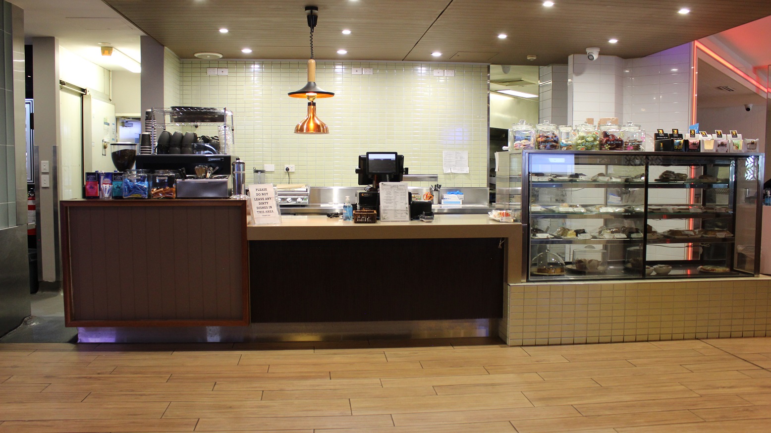 Another view of the café counter with a brown accents on the beige tiled walkway. The desk to the left have coffee packets, a large glass, a spoon and fork vessel, a flask, and a coffee cabinet with cups. The right, a food display shelf with cakes and snacks has snack jars and packers neatly organized on it. The checking screen, infographics, and sanitizers are on the middle. Behind are silver kitchen wares on a beige wall with an infographic and a mirror to the right. Another view of the café counter with a brown accents on the beige tiled walkway. The desk to the left have coffee packets, a large glass, a spoon and fork vessel, a flask, and a coffee cabinet with cups. The right, a food display shelf with cakes and snacks has snack jars and packers neatly organized on it. The checking screen, infographics, and sanitizers are on the middle. Behind are silver kitchen wares on a beige wall with an infographic and a mirror to the right.