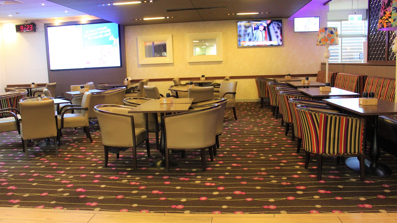 A brightly lit dining room with rows of chairs and tables. On the left, there are a few multicolored seats set horizontally against the wall, along with a large and small screen. Two heat stands are visible. In the centre, two rows of brown chairs and tables lead to a wall with a yellow frame pattern. The last row of multicolored chairs leads to the wall with a big screen. On the left and a timer. The floor is coated in a dark grey with a pink and yellow motif. A brightly lit dining room with rows of chairs and tables. On the left, there are a few multicolored seats set horizontally against the wall, along with a large and small screen. Two heat stands are visible. In the centre, two rows of brown chairs and tables lead to a wall with a yellow frame pattern. The last row of multicolored chairs leads to the wall with a big screen. On the left and a timer. The floor is coated in a dark grey with a pink and yellow motif.