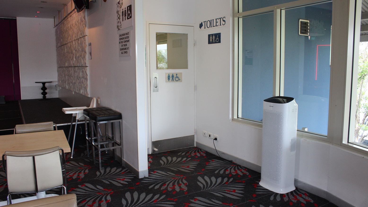 Behind a white marble wall with an infographic by the purple wall overlooking a space with a white highchair and two black high stools leading to the toilet. A white door with an infographic and a toilet tag on a grey and red covered floor surface, flanked by a white wall and a glass window on the left. A purifier can be seen standing by the wall on the right side. Behind a white marble wall with an infographic by the purple wall overlooking a space with a white highchair and two black high stools leading to the toilet. A white door with an infographic and a toilet tag on a grey and red covered floor surface, flanked by a white wall and a glass window on the left. A purifier can be seen standing by the wall on the right side.