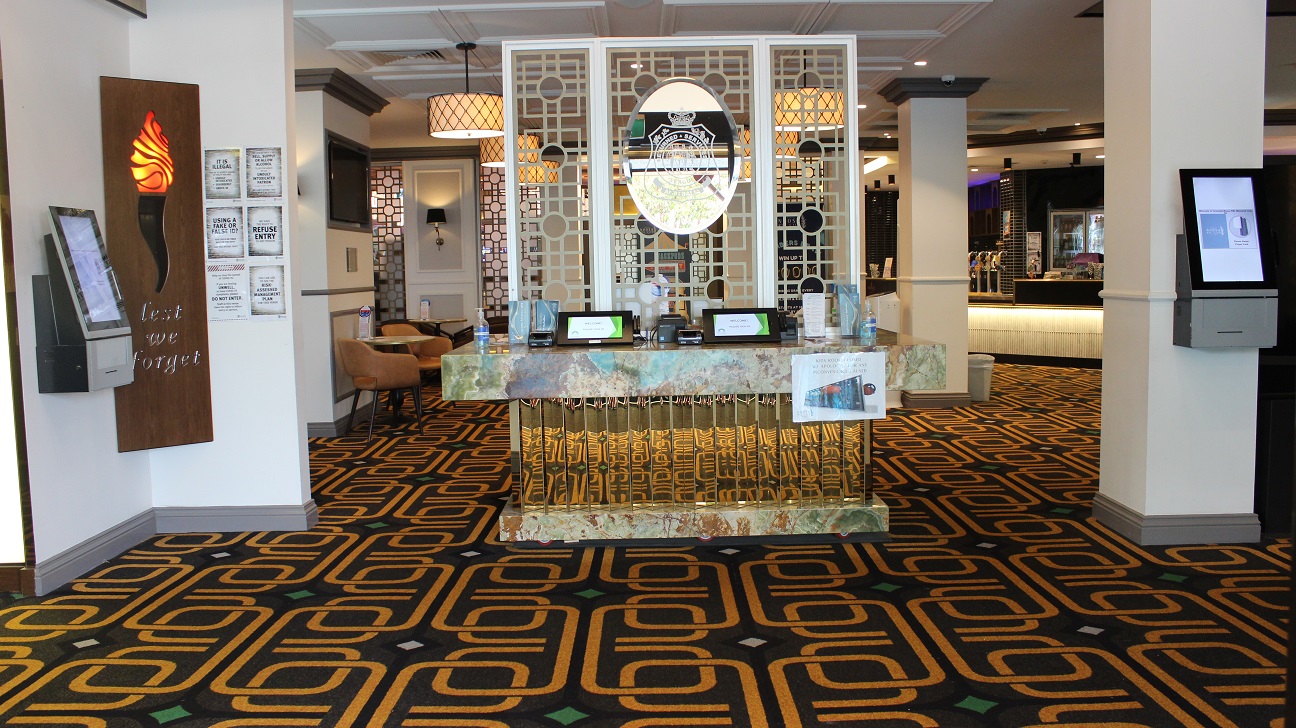 A table with a military emblem made of marble (white, brown, and vivid gold) stands in the centre of a foyer covered in brown and vibrant gold carpet and accents, barricading the seating space. Brown chairs, tables and a TV are on the left wall. Two pillars are visible, one with a check-in screen and the other with a rubbish bin. Ticket dispensers, infographics, and sanitizers, among other things, are on the table. At the far end, is the café. A table with a military emblem made of marble (white, brown, and vivid gold) stands in the centre of a foyer covered in brown and vibrant gold carpet and accents, barricading the seating space. Brown chairs, tables and a TV are on the left wall. Two pillars are visible, one with a check-in screen and the other with a rubbish bin. Ticket dispensers, infographics, and sanitizers, among other things, are on the table. At the far end, is the café.
