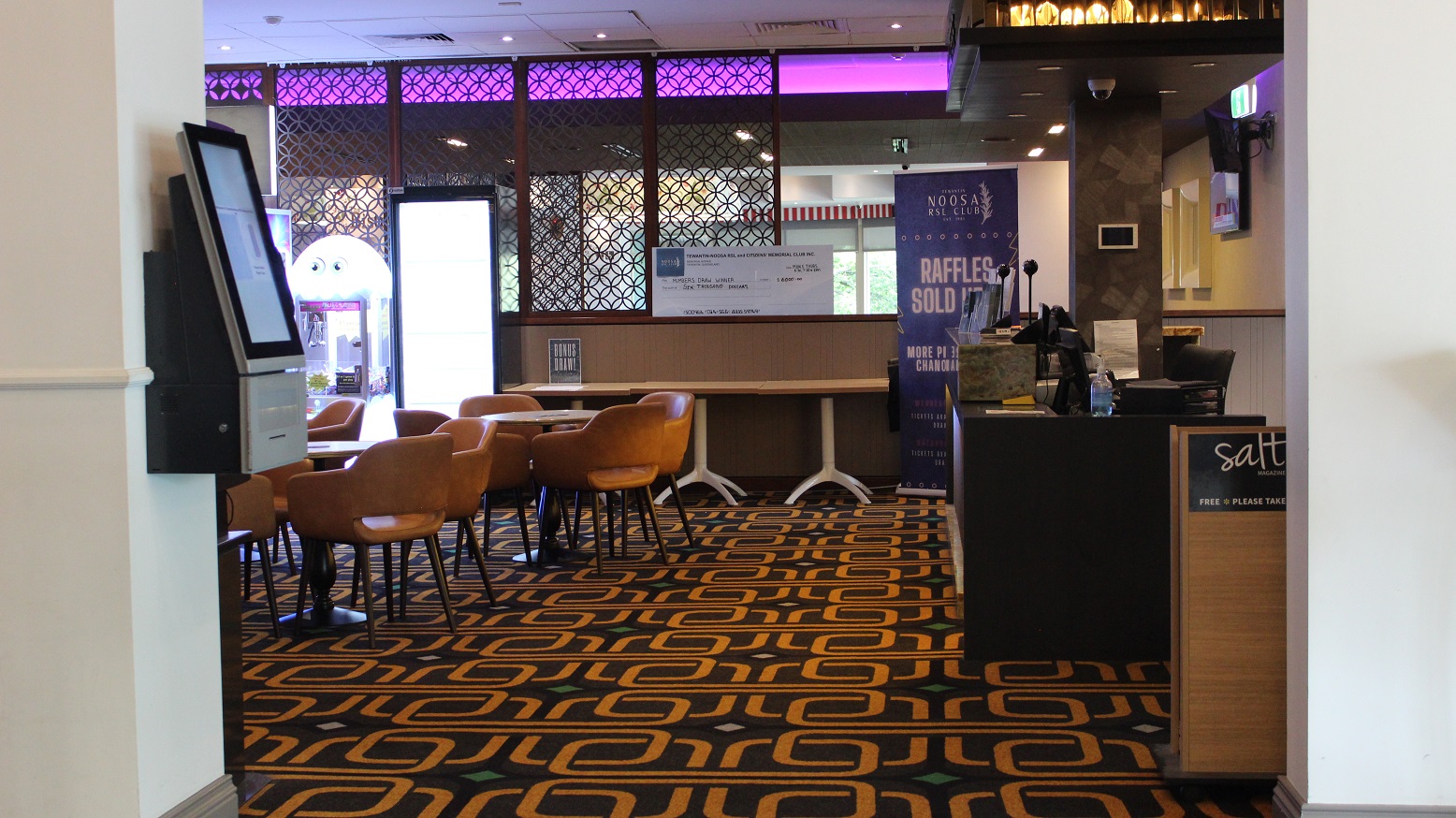 A seating space with brown chairs arranged around tables on a military-themed rug. A check-in device is on the right pillar. A demo cheque is set on the cubicle at the end with a brown barricade, which also has three tables in front by the Noosa raffle banner. A black checking desk has on it, two screens, books, sanitizer, and other materials, and a salt magazine box on the side. Behind the desk is a chair by a beige colour board with a screen. A seating space with brown chairs arranged around tables on a military-themed rug. A check-in device is on the right pillar. A demo cheque is set on the cubicle at the end with a brown barricade, which also has three tables in front by the Noosa raffle banner. A black checking desk has on it, two screens, books, sanitizer, and other materials, and a salt magazine box on the side. Behind the desk is a chair by a beige colour board with a screen.