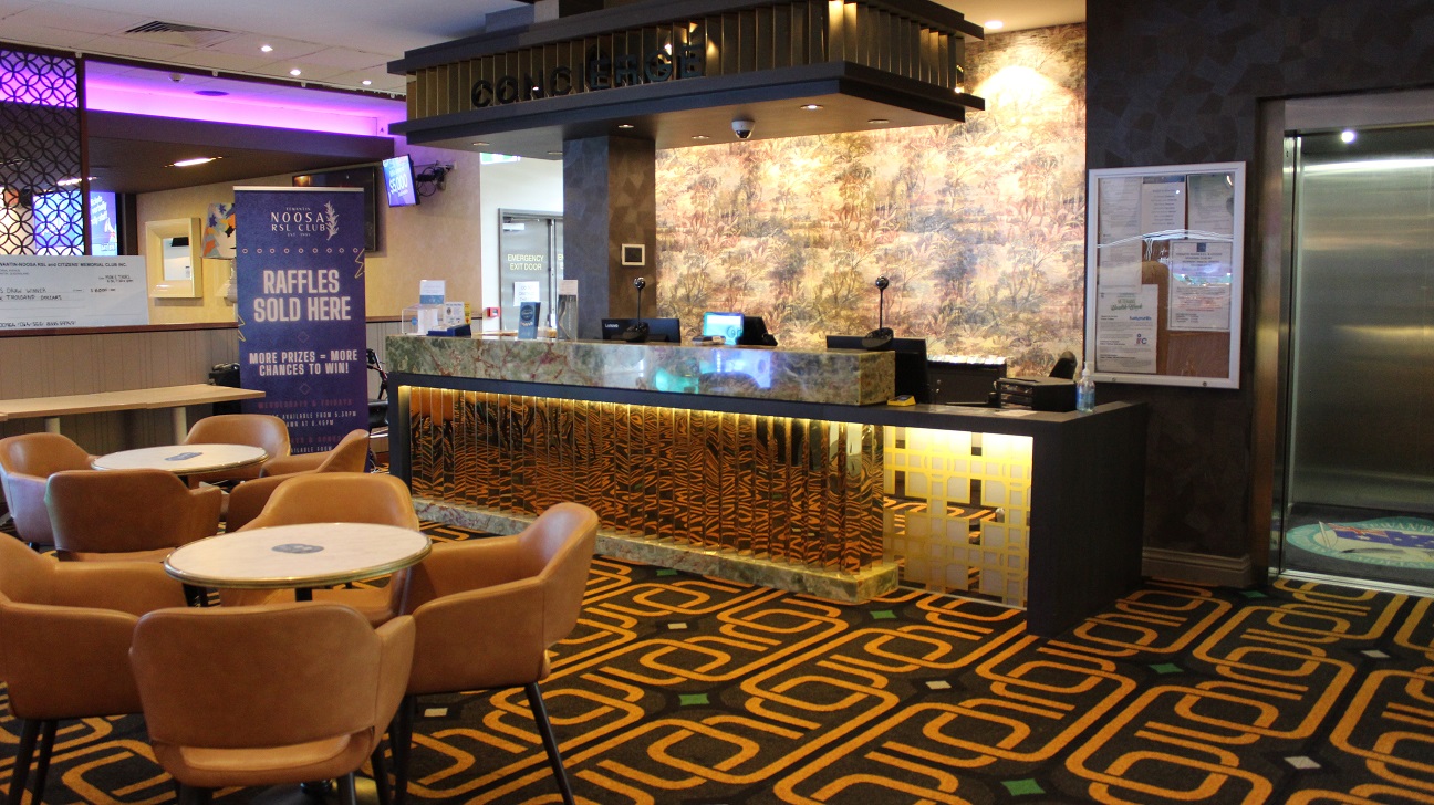 Another view of the previous image focusing on the well-lit spectacular counter decked out in strong gold and army accent. It has two upright speakers, four screens, boxes, sanitizers, and booklets. An emergency exit door with an infographic is on the left and the counter looks out onto the living room, which is furnished with two sets of four brown chairs arranged around a table. as well as a Noosa RSL club banner on the side. Another view of the previous image focusing on the well-lit spectacular counter decked out in strong gold and army accent. It has two upright speakers, four screens, boxes, sanitizers, and booklets. An emergency exit door with an infographic is on the left and the counter looks out onto the living room, which is furnished with two sets of four brown chairs arranged around a table. as well as a Noosa RSL club banner on the side.