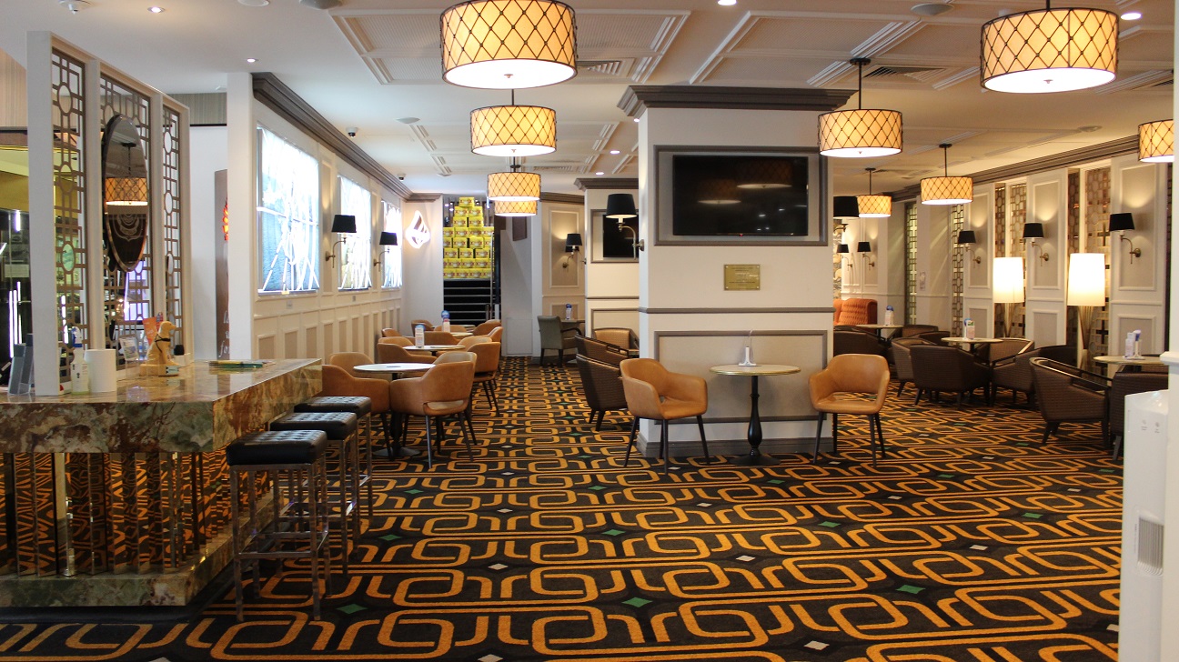 The dining area behind a table with the military motif emblem, and three stools resting on it. A napkin, ornaments, and other items are on the table. The well-lit area features brown chairs placed in rows around each table on a brown bright gold carpet. Chairs and tables lean against the two pillars, with TVs in the centre. At the other end, there are two orange seats. Every table has a menu in glass decor. The walls are white with brown accents and bright gold accents. The dining area behind a table with the military motif emblem, and three stools resting on it. A napkin, ornaments, and other items are on the table. The well-lit area features brown chairs placed in rows around each table on a brown bright gold carpet. Chairs and tables lean against the two pillars, with TVs in the centre. At the other end, there are two orange seats. Every table has a menu in glass decor. The walls are white with brown accents and bright gold accents.