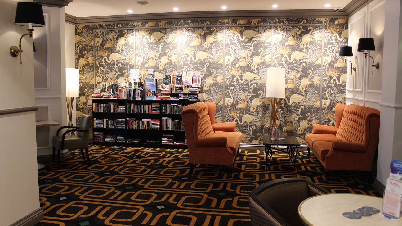 A view of the brown and yellow flamingo-themed wall, with a bookshelf to the left and two white-covered lamp fixtures. On the left are two sets of two cozy orange seats with a dark brown table in the centre. The walls are white, with a dark brown accent, and the floor covering is brown and gold. A view of the brown and yellow flamingo-themed wall, with a bookshelf to the left and two white-covered lamp fixtures. On the left are two sets of two cozy orange seats with a dark brown table in the centre. The walls are white, with a dark brown accent, and the floor covering is brown and gold.